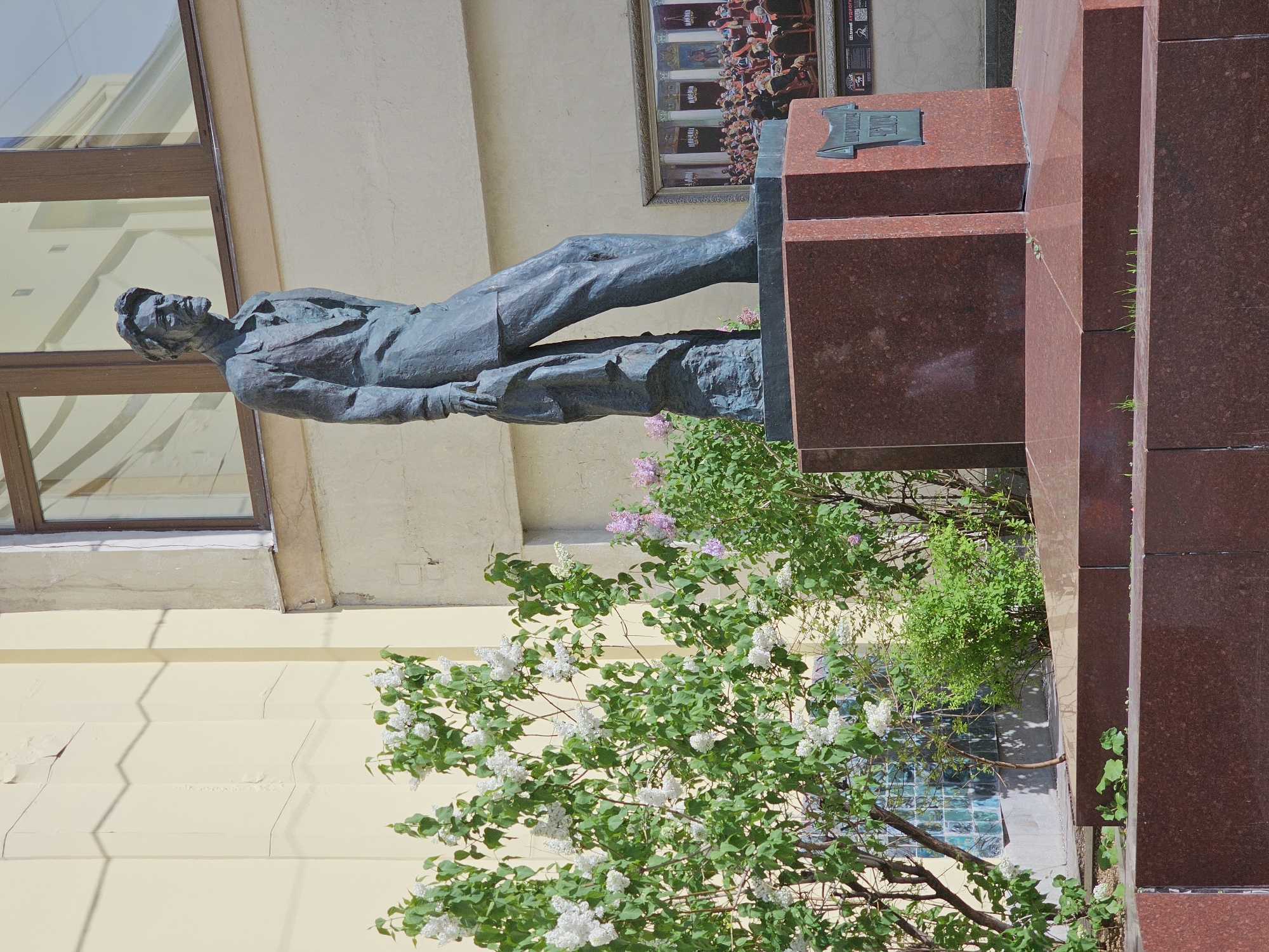 The image shows a statue of Sergey Yesenin, a Russian poet, in Moscow, Russia. The statue is bronze and stands on a red granite pedestal. White lilac bushes are blooming in front of the statue. The surrounding buildings appear to be institutional or residential, built of light-colored stone. A window of one of the buildings is visible behind the statue.