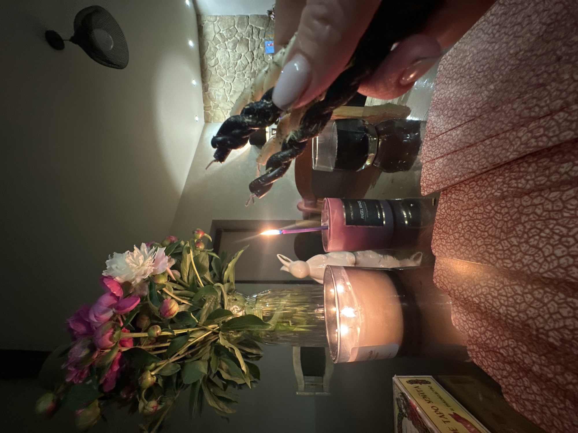 A close-up, low-angle shot captures a dimly lit tabletop scene. In the foreground, a fan of red-patterned cards lies spread out. To the left, a yellow and red box, identified as "DE TARO УЗИТА," is partially visible. Dominating the mid-ground are two lit candles: a large, pale orange candle in a glass jar and a smaller, purple candle with a lit wick. Behind these, a white ceramic bunny figurine stands. To the left of the candles, a clear glass vase holds a bouquet of peonies, with pink and white blooms and green foliage. In the background, a person's hand with manicured nails holds three dark, twisted objects, possibly incense sticks or dried herbs. A small glass of dark liquid sits on the table. The setting appears to be an indoor space with a stone-patterned wall and modern ceiling lights visible in the background, suggesting a home environment. There is no discernible indication of the city of Chaikovsky, Russia, within the image.