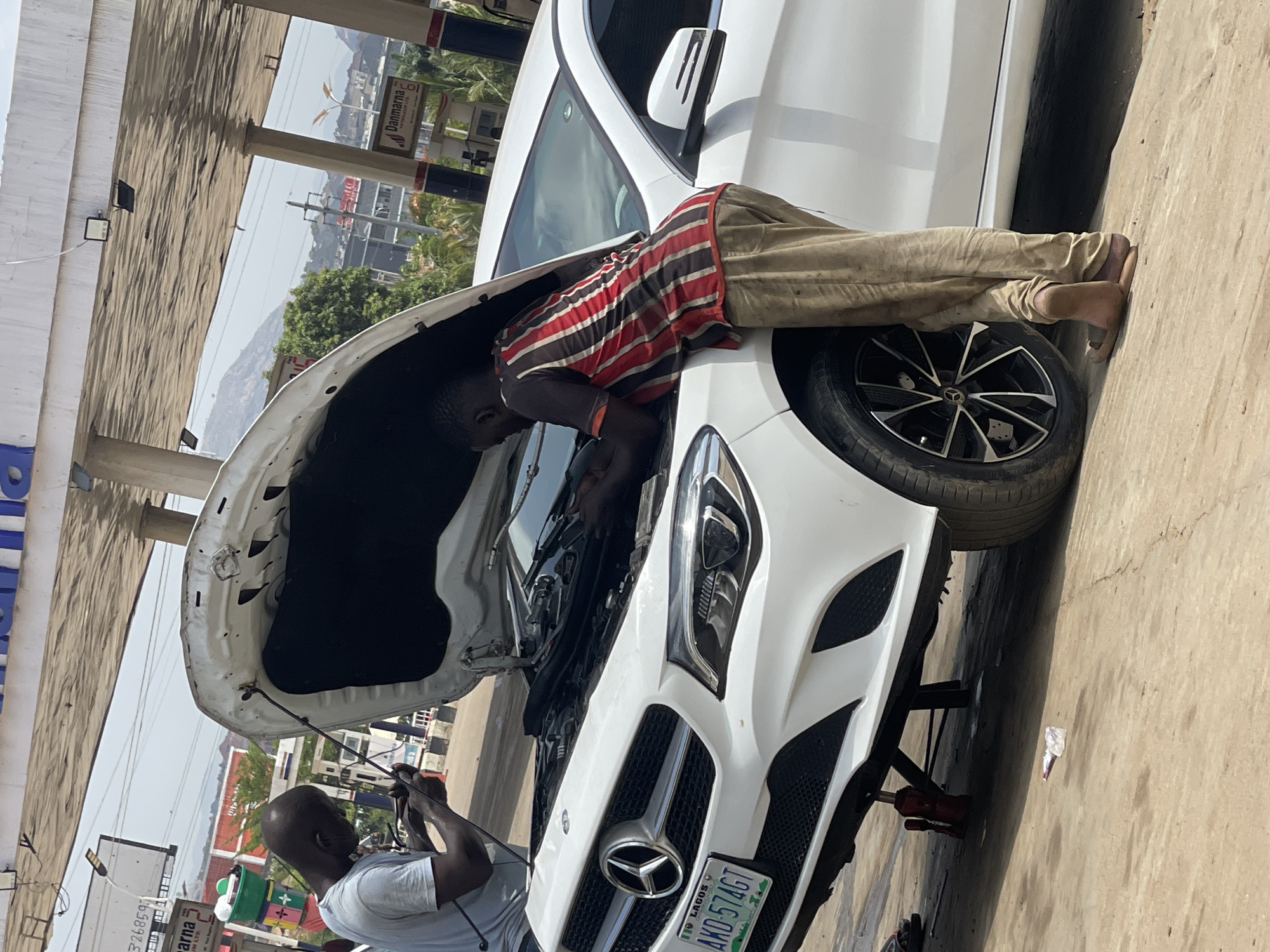 A mechanic is working on a white Mercedes car with the hood up at a gas station in Galadimawa Village, Nigeria. The mechanic, wearing a striped shirt and tan pants, is leaning into the engine compartment. Another person is visible near the front of the car. The car is supported by a jack. The scene appears to be a daytime outdoor setting. A sign mentioning "Danamarna" and what might be a gas price is visible.