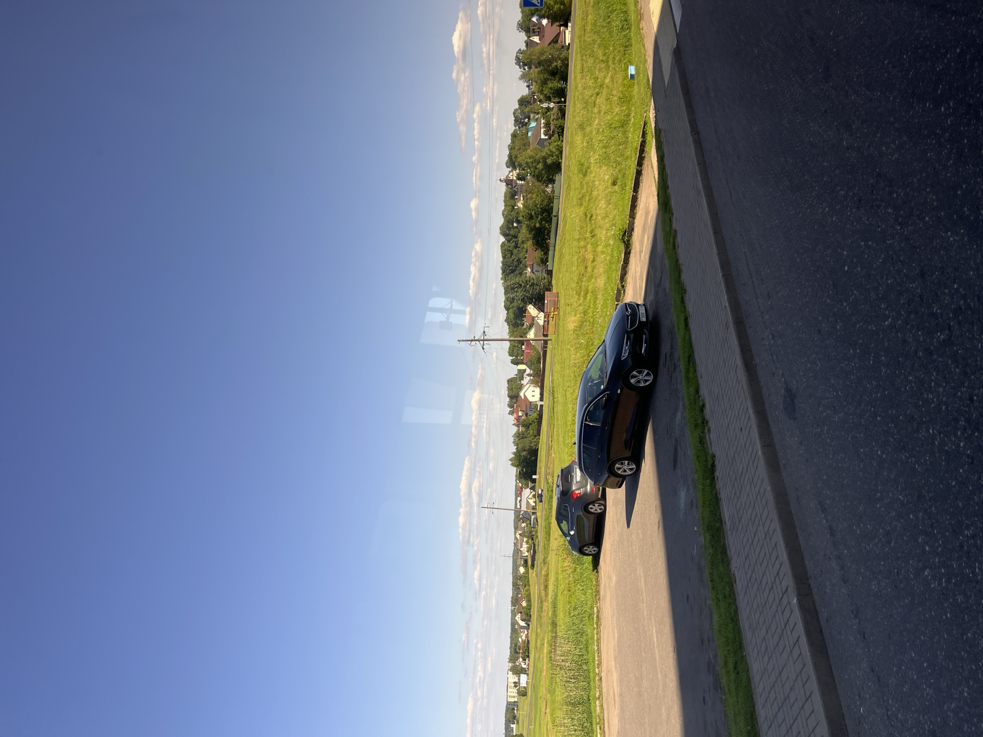 In Myadzyel, Belarus, a sunny day presents a landscape view from a paved area. Two cars are parked on a gravel lot adjacent to a grassy field. The field slopes upwards towards a treeline dotted with houses and a church spire. The sky is a clear, bright blue with scattered white clouds. A reflection of what appears to be a window frame is visible in the upper part of the image.