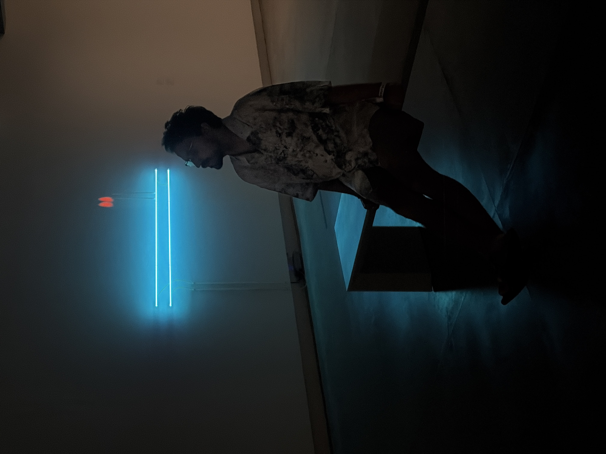 The photograph shows a person standing in an art gallery. The person is a man, with short dark hair and glasses, wearing a light-colored patterned shirt and dark shorts. He is looking down at a piece of artwork, which is a blue neon sculpture consisting of two vertical lines and a horizontal arrow shape to the left. The artwork is mounted on a white wall. The person is standing on a wooden platform that is casting a shadow. The overall scene is dimly lit, with the blue neon sculpture being the primary source of light. The background is dark, suggesting the setting is an indoor space, most likely an art gallery. Bangkok, Thailand is not discernible in this image.