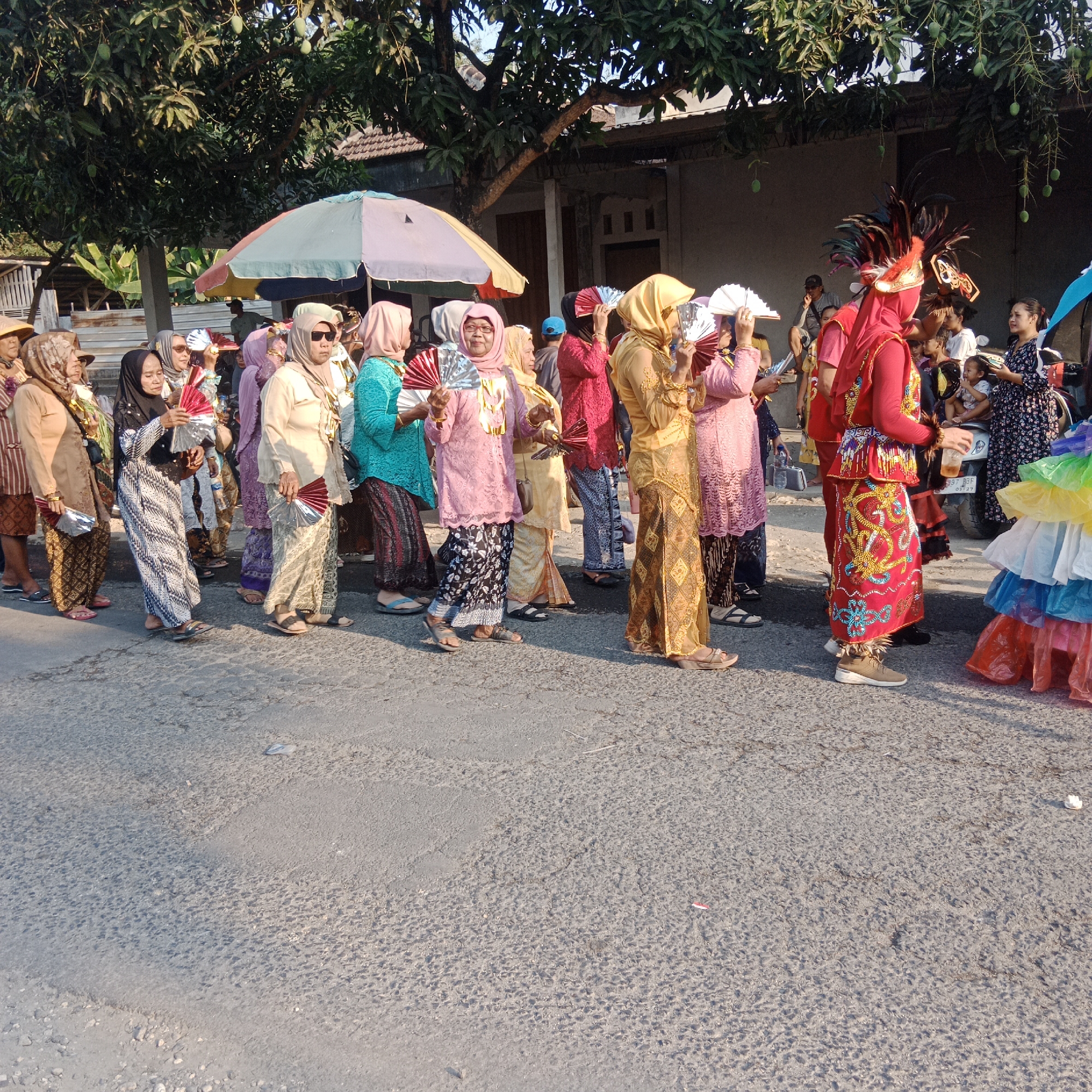 The image depicts a procession or festival taking place on a paved street in Tegalrejo, Indonesia. A group of people, mostly women, are walking in a line, dressed in traditional attire. Many are wearing headscarves and colorful clothing, with some holding fans. One individual in the center wears a prominent headdress with feathers and a red, ornate outfit, suggesting a ceremonial role. The background shows trees with fruit, a building, and a street umbrella. The lighting indicates it is daytime, possibly late afternoon due to the long shadows. The ground surface is asphalt with some gravel.
