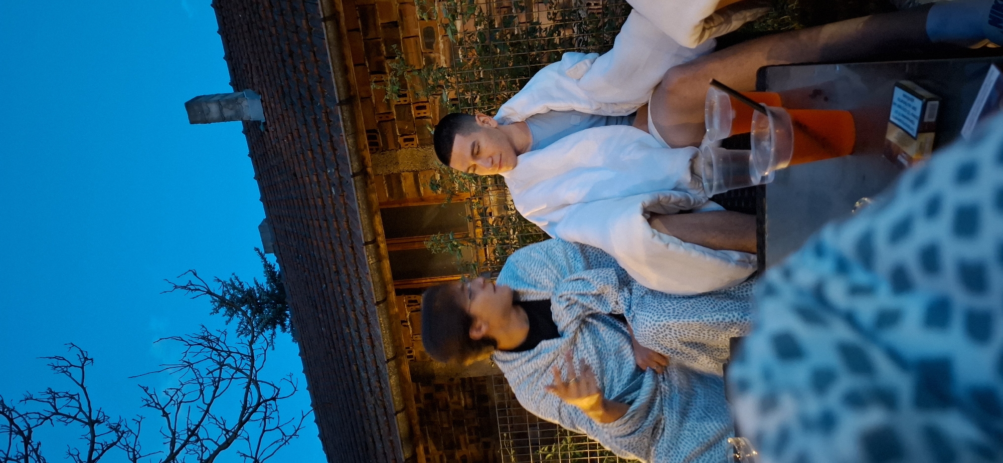 The image depicts two young men sitting outdoors in the evening, wrapped in blankets, with a building in the background. The man on the left is wearing glasses and a black t-shirt, with a patterned blanket draped over him. He is gesturing with his left hand, as if speaking. The man on the right is wearing a light blue t-shirt, with a white blanket covering him. He is looking to his right, with a neutral expression.
In front of them, on a table, are several plastic cups, some containing red liquid and straws, and a pack of cigarettes. The building in the background appears to be made of brick with a tiled roof and a chimney. A bare tree branch is visible in the upper left corner of the frame against a deep blue sky.
The scene suggests a casual gathering in an outdoor setting. The time of day is dusk or early evening, indicated by the twilight sky. The location is the city of Lobosh, Bulgaria.