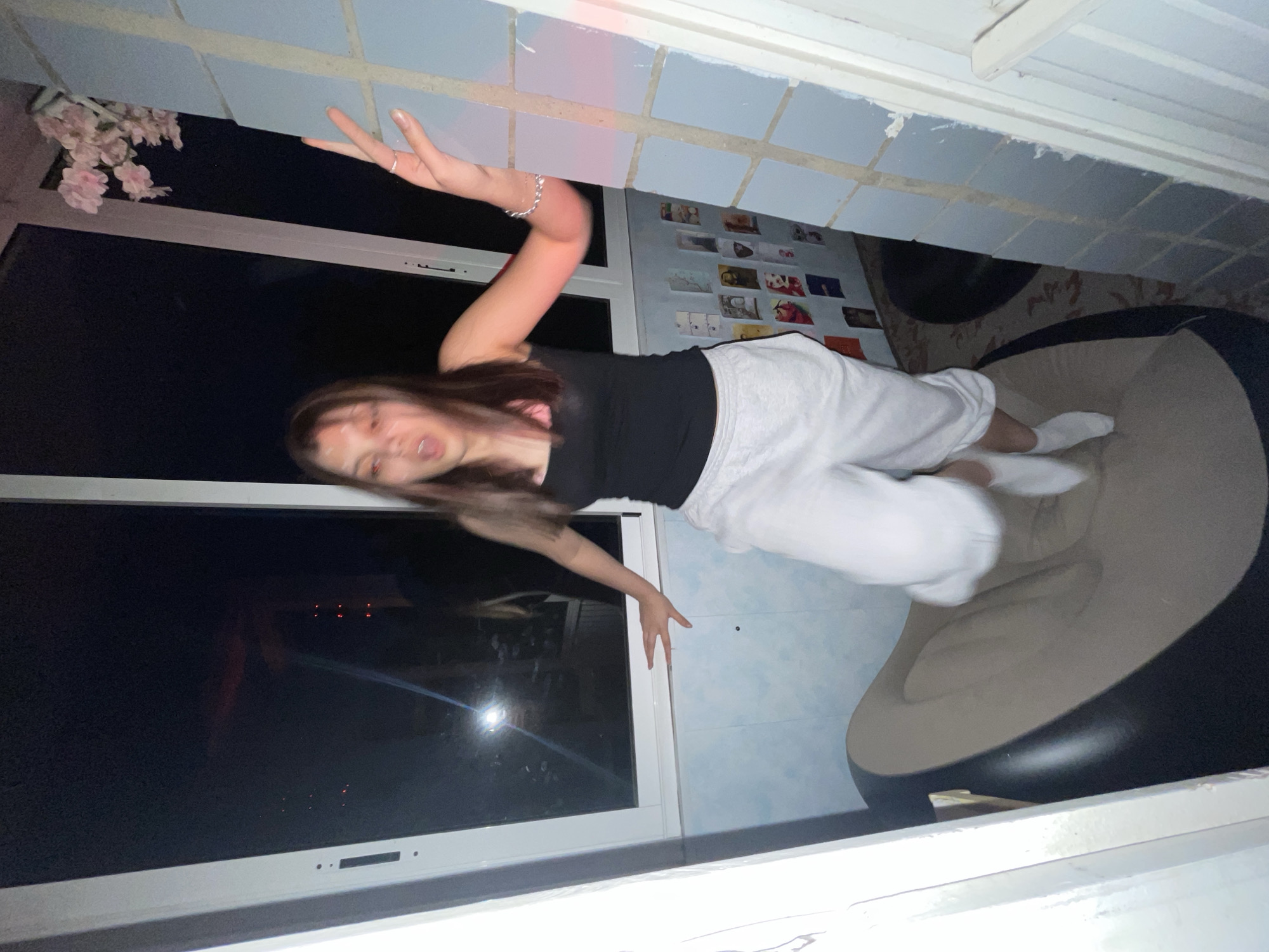 The image, rotated 90 degrees counter-clockwise, depicts a young woman in energetic motion within an indoor setting at night.
The central subject is a young woman with long, dark hair, which is visibly blurred, indicating rapid movement. She is wearing a black short-sleeved top, light grey sweatpants with a dark stripe, and white socks. Her face is also blurred, with her mouth slightly open, suggesting an active expression. Her arms are outstretched; one hand reaches towards a tiled surface, and the other is extended outward. A silver chain bracelet adorns her right wrist.
She appears to be mid-air, possibly jumping or falling onto a large, two-toned inflatable chair or beanbag, which occupies the lower right portion of the frame. The chair has a black base and a lighter, beige-colored top.
The background consists of light blue tiled walls or ceiling sections. To the left, a large window reveals a dark exterior, indicating nighttime. Reflections of distant lights, appearing as small red dots, are visible on the windowpane. A white window frame borders the glass. In the upper right background, a wall is decorated with numerous small, casually arranged photographs or postcards. A small branch with pink, cherry-blossom-like flowers is visible near the top left corner, adding a decorative touch.
The scene takes place indoors, likely a room or an enclosed balcony, at night. The activity suggests a playful or candid moment, characterized by the woman's dynamic pose and the motion blur. No discernible text is visible.