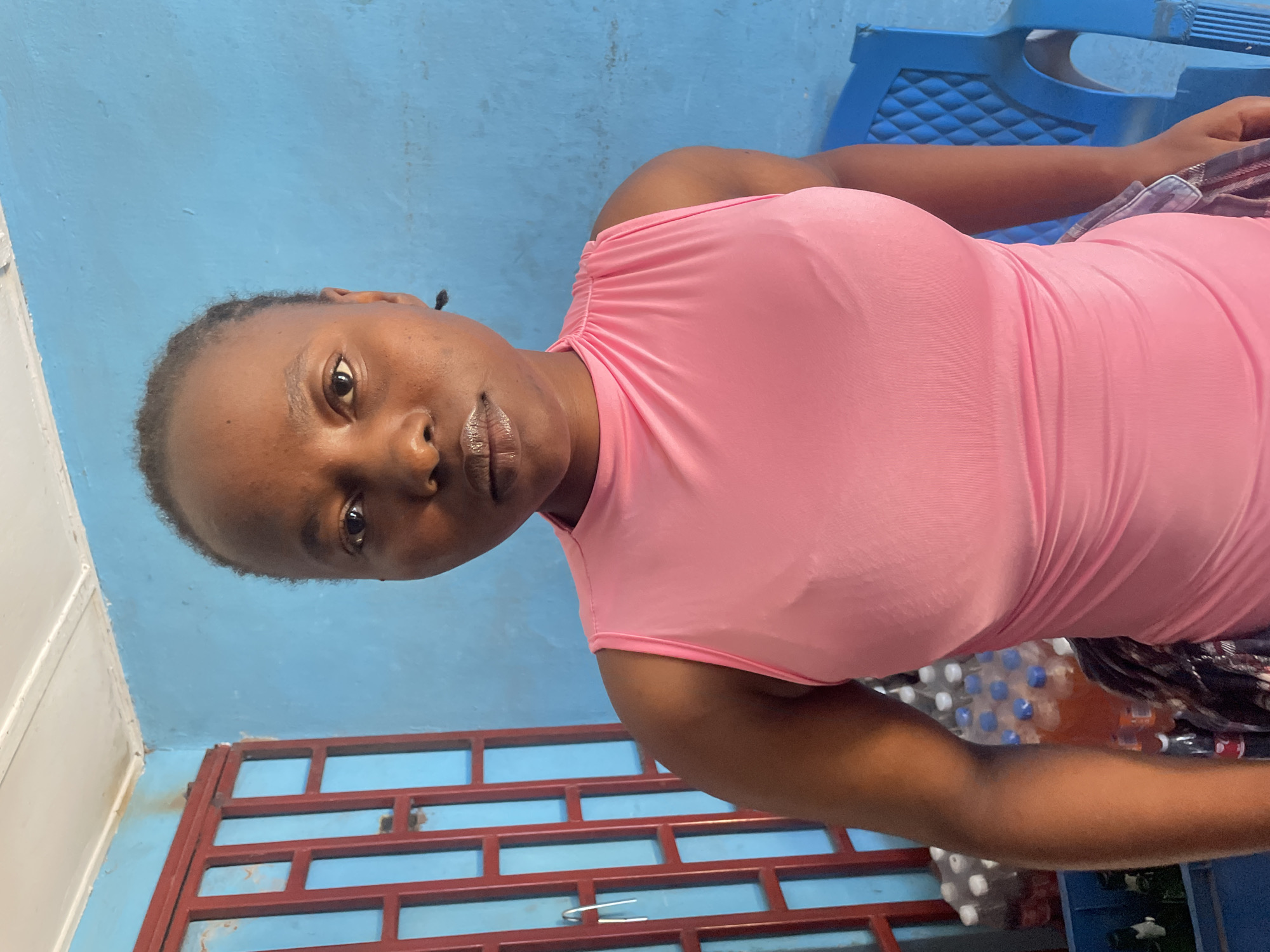 A woman wearing a pink top is seated outdoors. A blue plastic chair is behind her. A red metal grate is in the foreground. Bottles of water are next to her. The scene is in Jos, Nigeria.