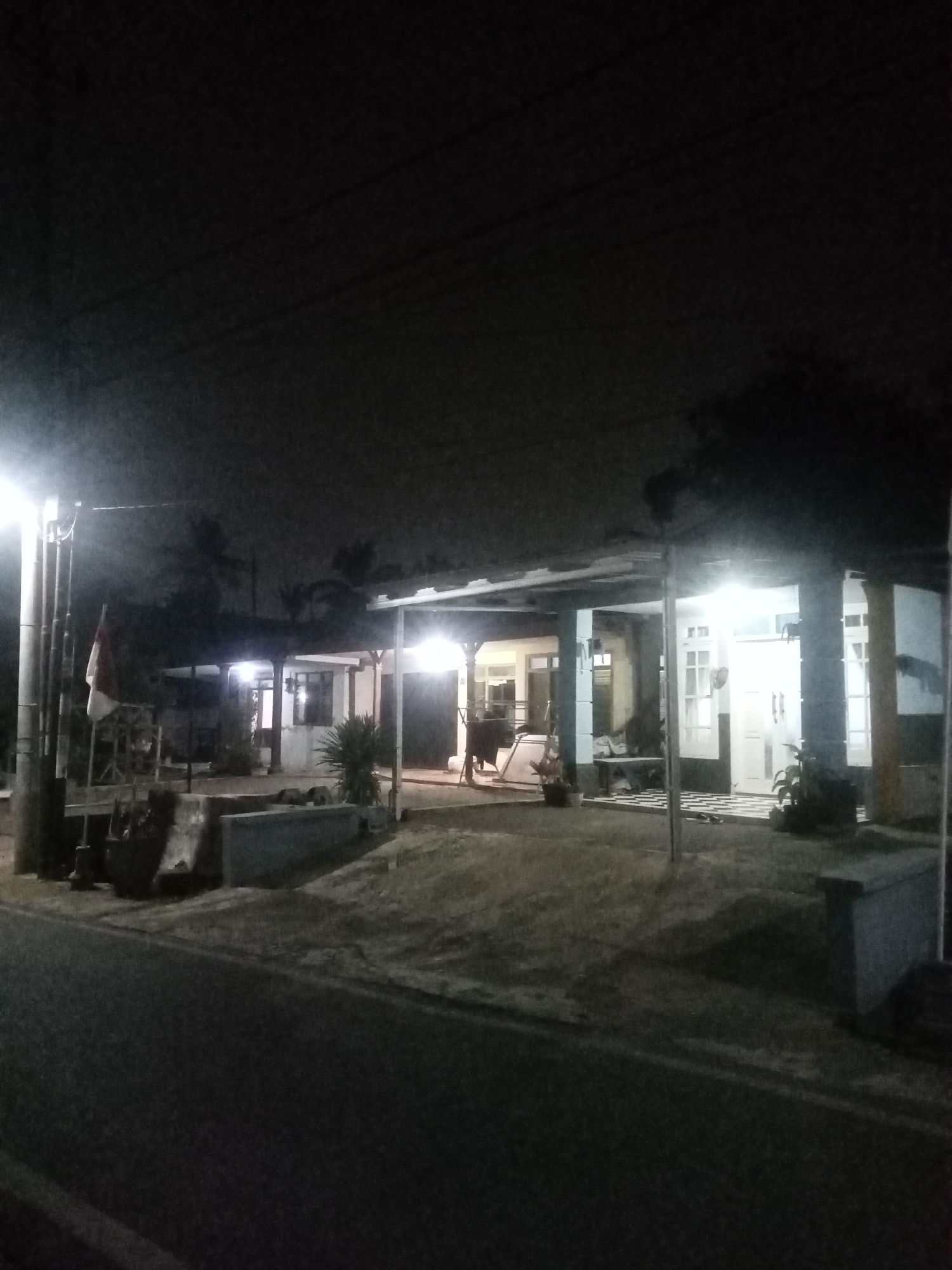 The image depicts a street-level view of a building with a porch, illuminated by artificial lights at night. The building appears to be residential or a small commercial establishment. A flag, which appears to be the Indonesian flag (red and white), is visible on a flagpole to the left of the building. There are trees and vegetation around the building. The road in the foreground is asphalt. The scene suggests a quiet street in Malang, Indonesia, given the presence of the Indonesian flag and tropical vegetation. No people are clearly visible in the image. The overall atmosphere is dark, indicating nighttime, with streetlights and interior lights providing the illumination.