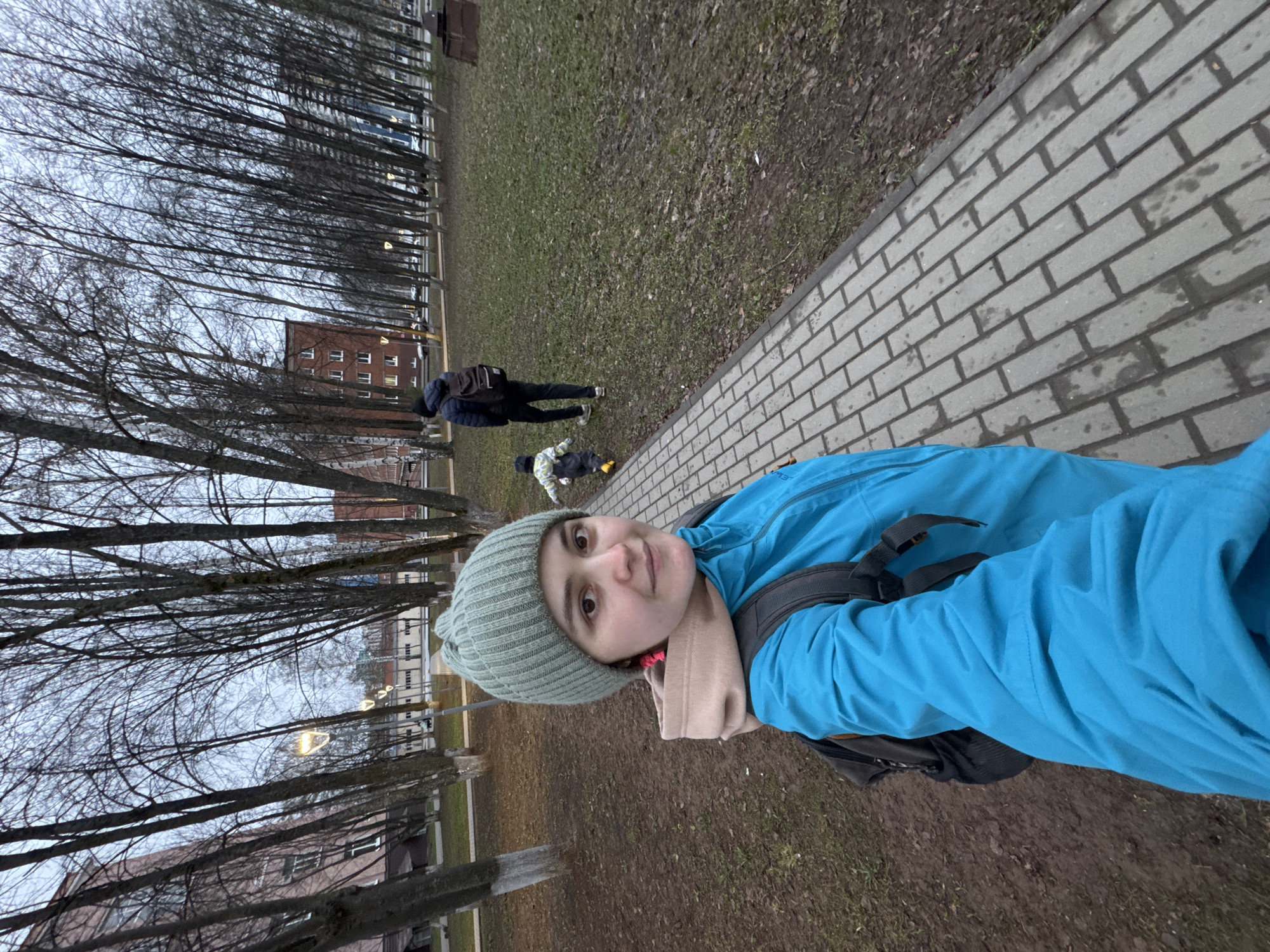 The image shows a person, possibly a young woman, taking a selfie on a paved pathway adjacent to a grassy area. The woman wears a light blue jacket, a gray-green knit hat, and a tan scarf. She has a backpack on. In the background, two other individuals are visible: an adult wearing a dark jacket and carrying a backpack walking and a child in a light-colored jacket lying down. The environment includes a row of trees along the pathway and buildings in the distance. Based on visual cues such as the architecture and bare trees, it might be autumn or early spring. The location is speculated to be in Izhevsk, Russia but without further evidence, this remains a speculation.