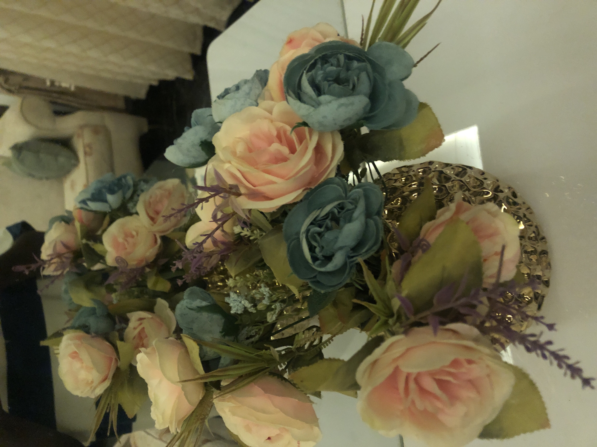 A close-up shot displays a bouquet of artificial roses and lavender in a textured, gold-colored vase. The flowers are a mix of soft pink and teal hues, with green foliage. The vase is positioned on a white surface, likely a table, and the background is out of focus, showing hints of furniture and drapery. The lighting in the scene appears to be artificial, casting a soft glow on the flowers and vase, with a bright reflection on the white surface. There are no people visible in the image. The scene does not provide any specific context or clues about its location within Bar Jirgi Summa, Nigeria.