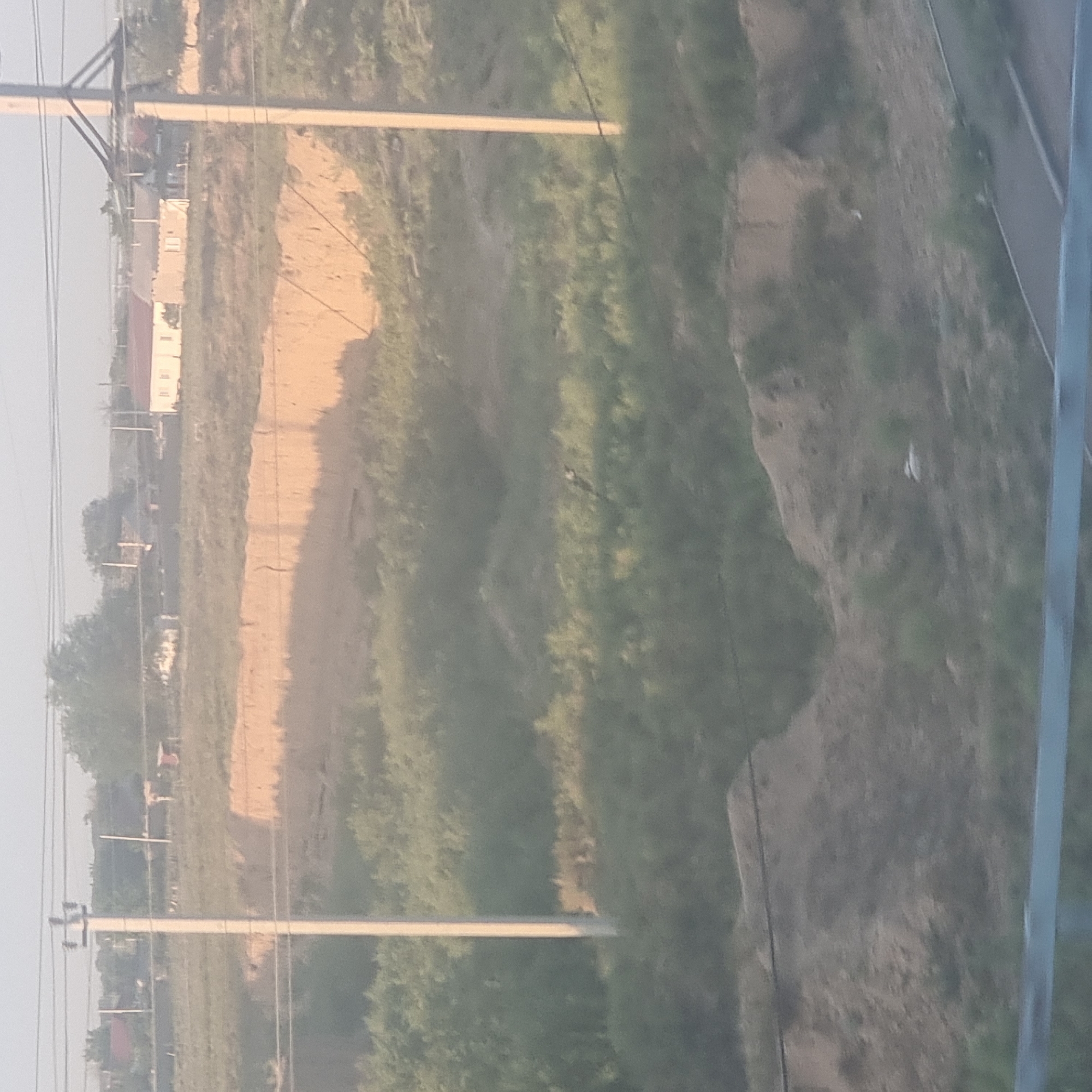 The image captures a landscape near the city of Arys, Kazakhstan. In the foreground, a blurred metal barrier is visible at the bottom. Behind it, a sloped, earthy embankment covered in sparse vegetation dominates the lower half of the image. A distinct, lighter-colored, eroded cliff face with horizontal striations is visible in the middle ground, suggesting a change in terrain or a man-made excavation. Further back, more vegetation, primarily green shrubs and trees, covers the undulating landscape. In the distance, several buildings, including houses with red and light-colored roofs, are scattered across the horizon. Two utility poles with wires are prominent features in the scene, one in the center and another to the right, indicating the presence of infrastructure. The sky is a pale, hazy blue, suggesting a bright but possibly overcast or dusty day. The overall impression is a rural or semi-rural setting with evidence of both natural and human influence on the environment.