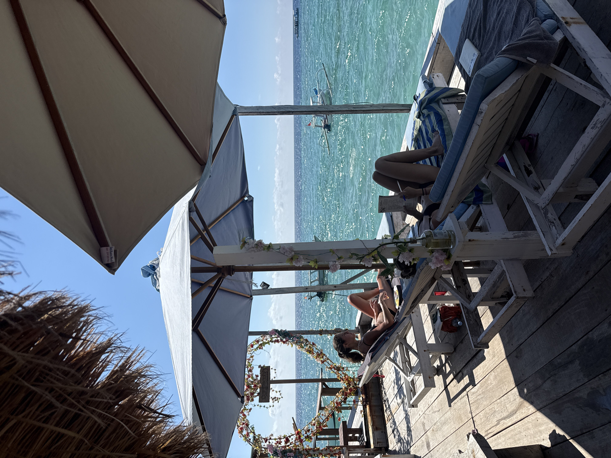 The image captures a seaside scene, likely in Unknown, Indonesia. The foreground features a wooden deck furnished with white-painted lounge chairs, one occupied by a woman in a bikini reclining and using a phone. A heart-shaped floral decoration adorns the deck's edge, accompanied by sun umbrellas providing shade.
The middle ground shows a turquoise sea with a small boat in the distance. The background reveals a clear, blue sky meeting the sea at the horizon. On another lounge chair, the lower body of a person is visible, with a book or menu, and writing utensil. The scene suggests a relaxing, beachside setting.