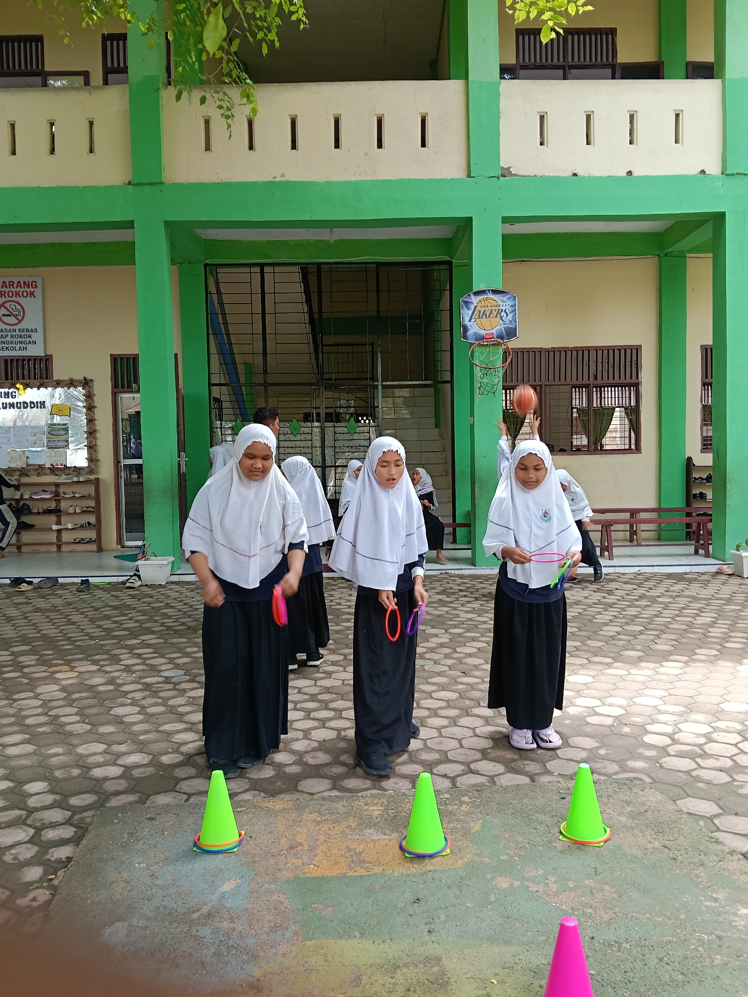 In Lhokseumawe, Indonesia, a group of students wearing white headscarves and black skirts are engaged in a ring toss game outdoors. They are positioned near green and pink cones. In the background, a green and beige building features a basketball hoop with a "Los Angeles Lakers" logo. A bulletin board and a shoe rack are visible near the building's entrance, while a basketball game appears to be in progress on the basketball court in the background.