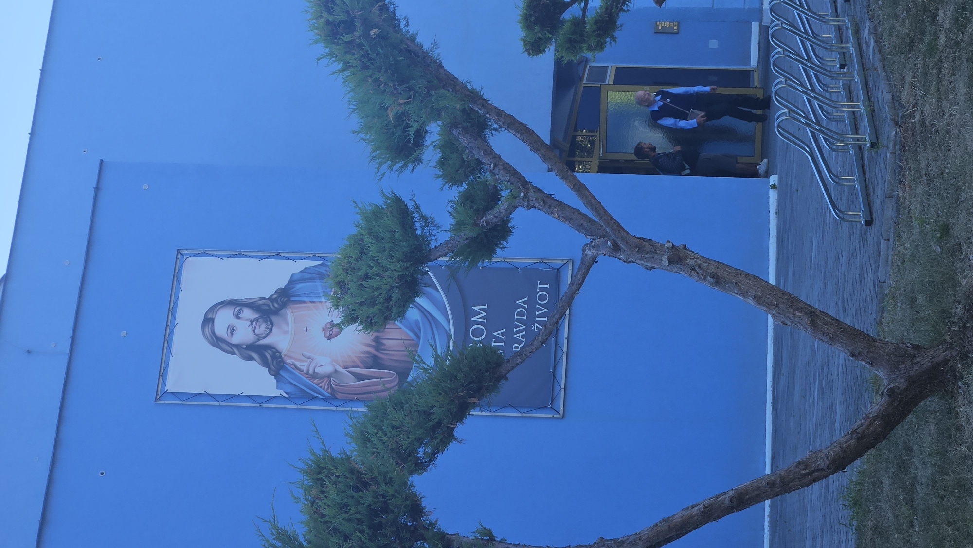 In Prešov, Slovakia, an outdoor scene features a bright blue building with a large banner depicting Jesus Christ with his hand over his heart. Below this image, a smaller banner displays text, with the visible words being "KOM", "TA", "RAVDA", and "ŽIVOT". In front of the building, two trees with sparse foliage frame the view. To the right, an entranceway is visible, with two men standing near it. One man, wearing a blue shirt and a dark vest, is walking away from the entrance, while another man, with a backpack, stands closer to the doorway. In the foreground, a bicycle rack with multiple slots is positioned on a paved surface. The overall lighting suggests it is daytime, with a slightly overcast sky.