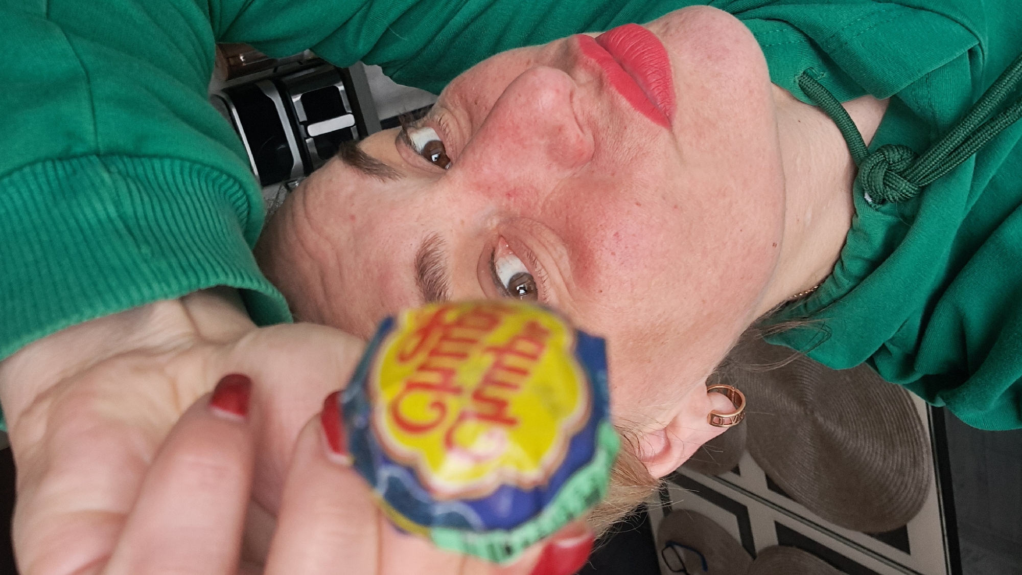 A woman in a green hooded sweatshirt is holding a Chupa Chups lollipop. She has red lipstick and red fingernails. She appears to be indoors with a patterned floor visible in the background. The image does not provide enough information to verify the location as Elektrostal, Russia.