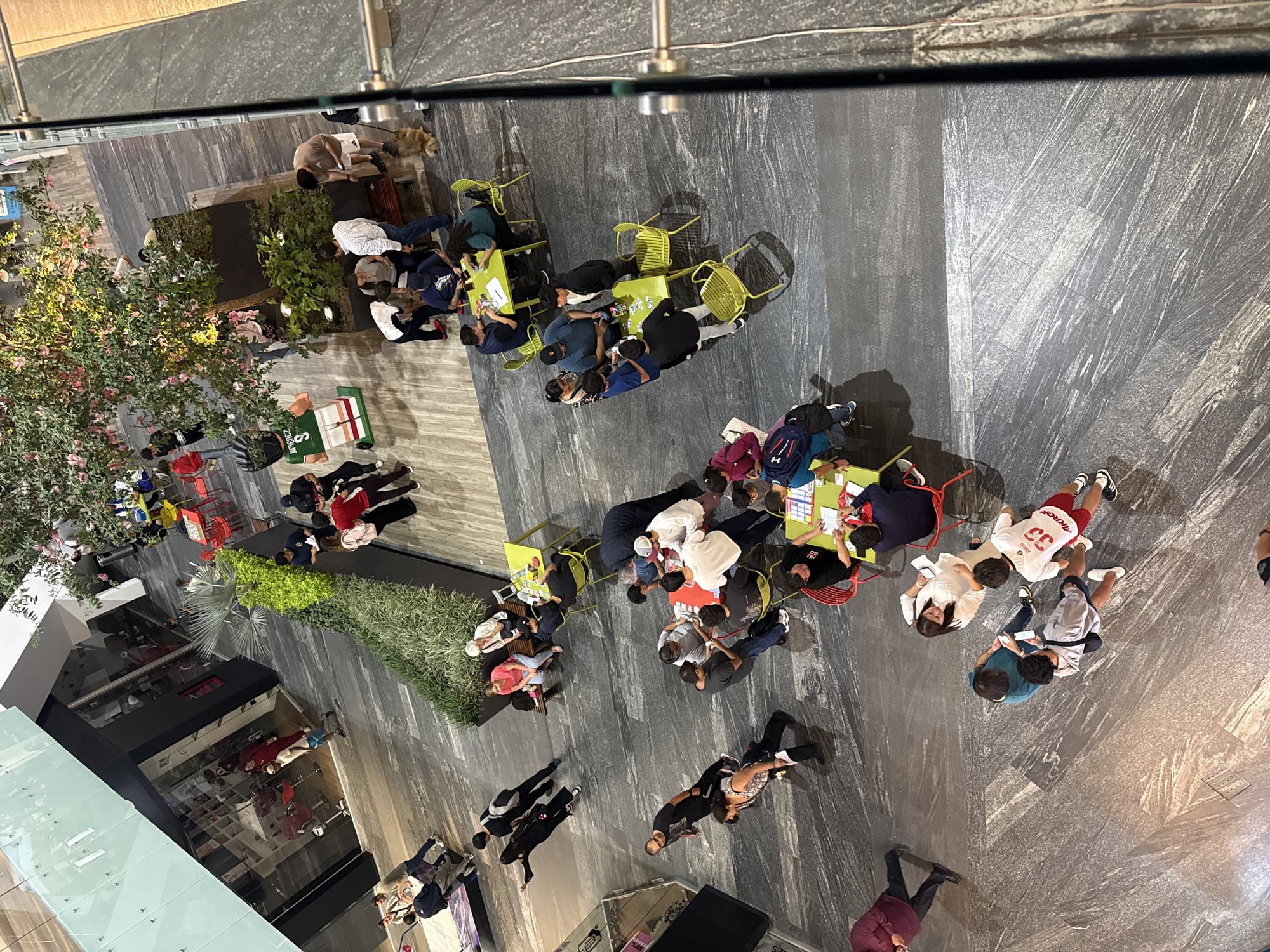 This aerial photograph captures a public space in Puebla, Mexico, featuring multiple groups of people engaged in various activities. The location appears to be an outdoor section of a shopping center or similar commercial area, as suggested by the presence of storefronts, glass partitions, and constructed landscaping.
Several clusters of people are seated at tables, some of which are yellow and red. The people are engaged in social interaction, such as dining, playing games, or casual conversation. Some individuals are standing or walking through the area.
The environment includes tiled or paved flooring, which has a grey color. The lighting suggests that the photograph was taken during the day, or at night with artificial lighting. There are planted trees and shrubs, adding a touch of greenery to the hardscaped environment.