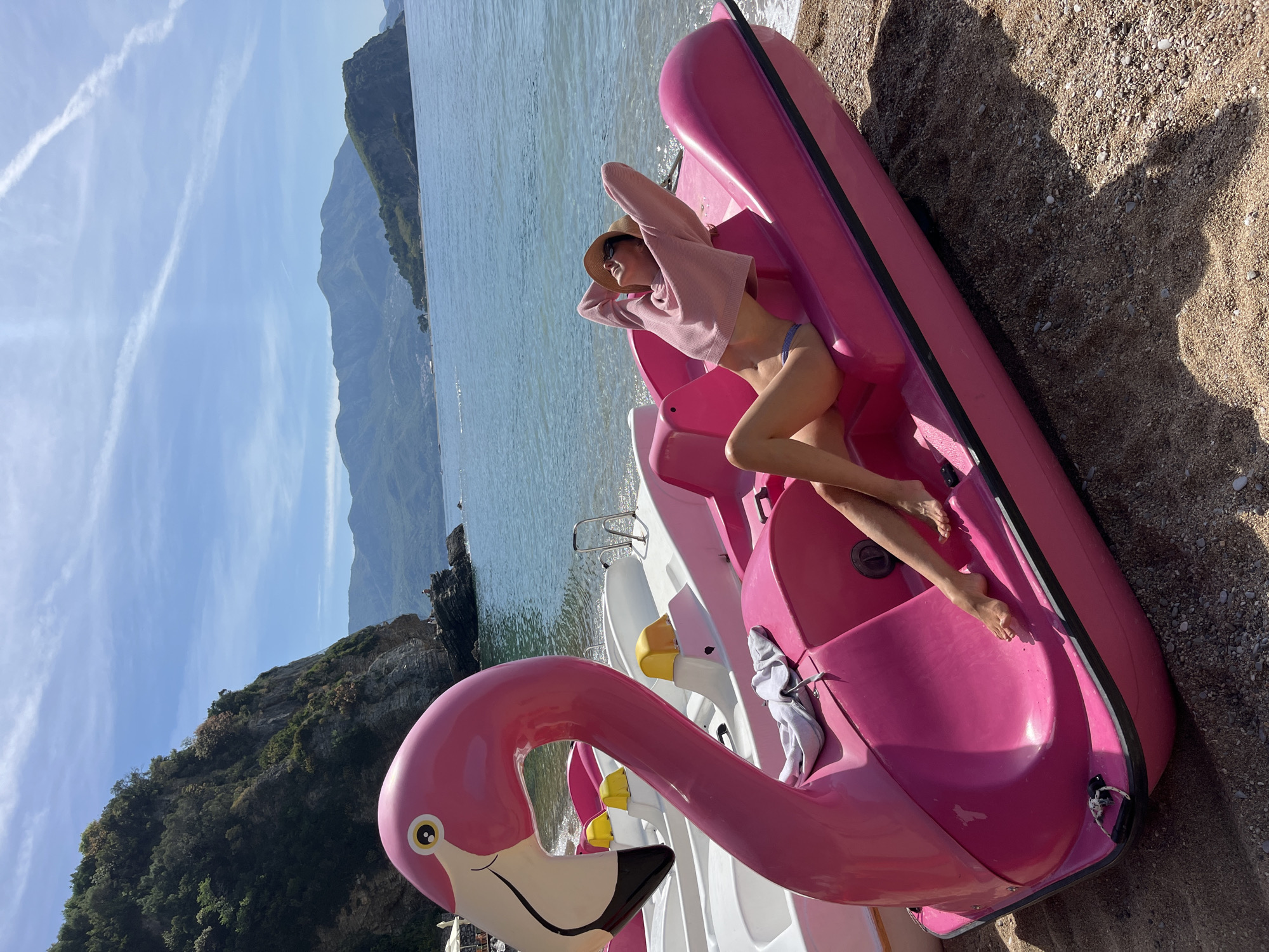 A woman is lying in a pink, flamingo-shaped paddle boat on a beach in Budva, Montenegro. She is wearing a pink sweater, blue bikini bottoms, and a straw hat. Her hands are behind her head. The paddle boat is on a sandy beach, and the sea is in the background. There are mountains in the distance and clear blue sky.