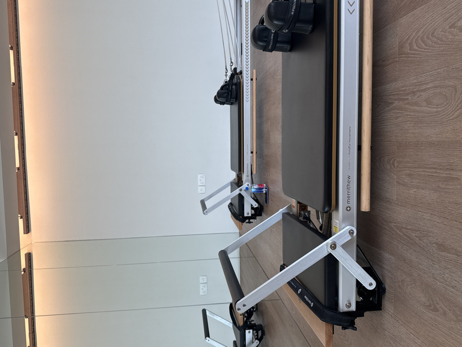 A Pilates studio located in Dubai, United Arab Emirates, is depicted. Two Merrithew brand Pilates reformer machines are the central focus. The foreground machine, partially cropped, features black padding, a light wooden frame, and silver-colored metal components. Behind it, a second Merrithew reformer is fully visible, sharing the same black padding, wooden frame, and silver-colored parts. A small blue plastic water bottle, identifiable as Fiji brand, rests on the wooden base of the second reformer near its foot bar. The studio floor is light brown, with a wood-grain laminate finish. The walls are light grey, and large mirrors line the left side of the room, reflecting the reformers and two white wall-mounted electrical outlets, each with two sockets. Above the mirrored section, a linear wooden fixture with integrated LED lighting is installed, emitting a warm glow onto the wall. No individuals are present in the scene, and no activity is observed.
