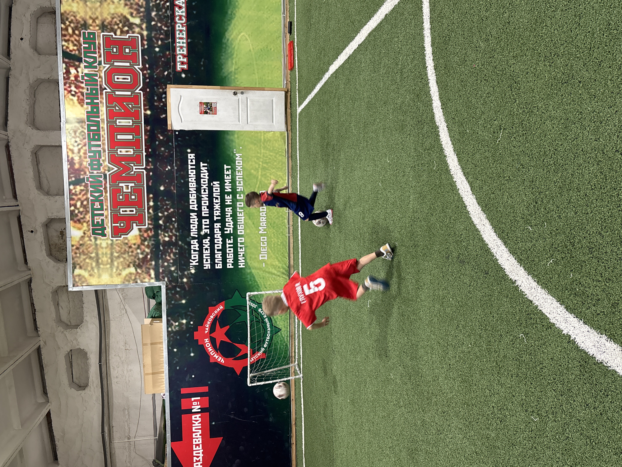 The image shows an indoor soccer field. Two children are playing soccer, one wearing a red jersey with the number 5, the other a blue jersey with the number 8. A soccer ball is near the goal. In the background is a banner for a children's soccer club called "Champion", located in the city of Chaikovsky, Russia. The banner features the club's logo, a quote from Diego Maradona, and the word "Coach".