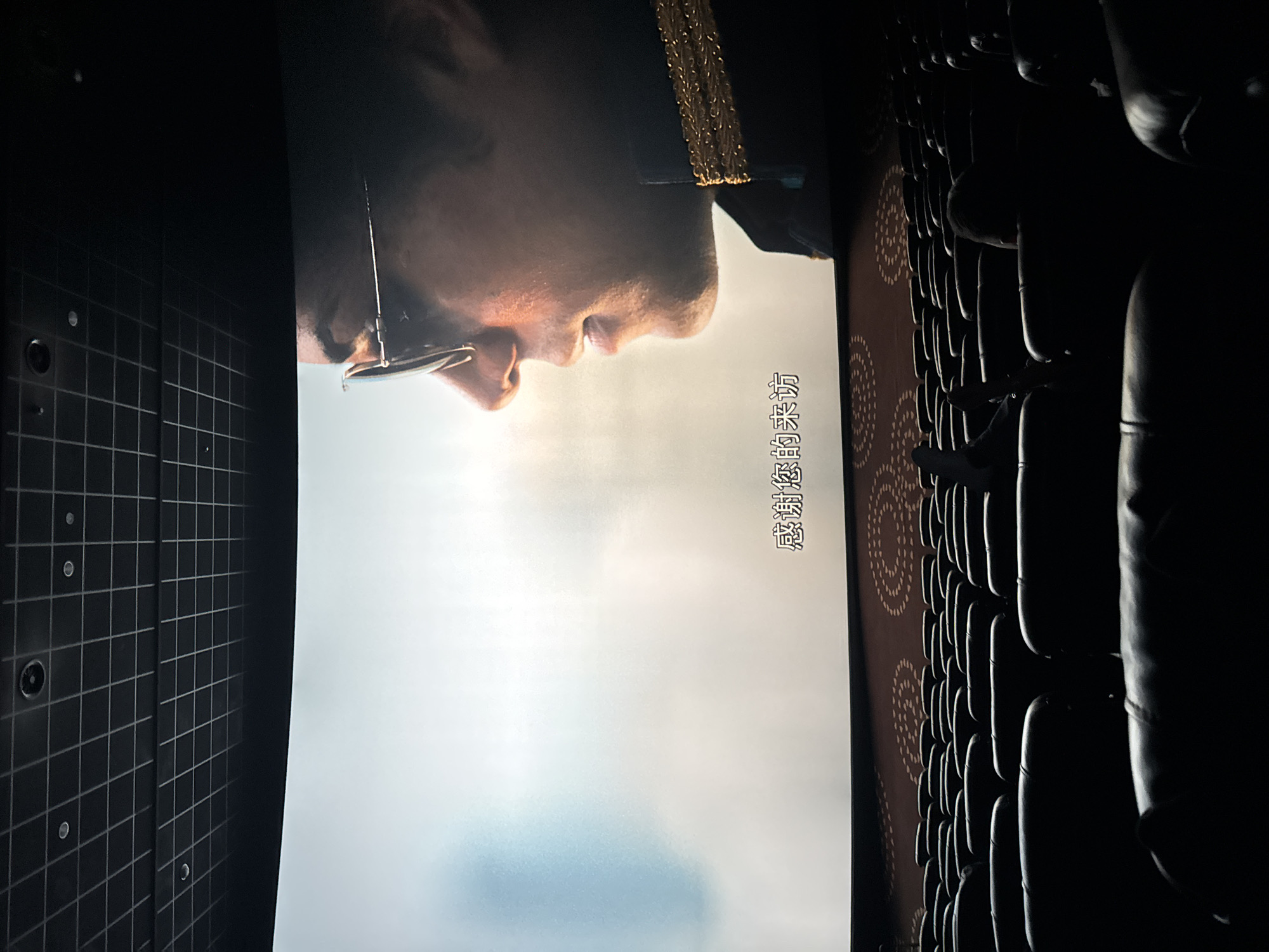 The image shows a view from a movie theater in Taihe District, China. On the screen is a close-up of a man's face, likely an actor in the film. He is wearing glasses and a dark uniform with gold detailing at the collar. The Chinese characters "感谢您的来访" are visible on the screen, which translates to "Thank you for your visit." The theater itself is dark, with rows of empty seats in the foreground and to the side of the screen. The left wall is visible, with a grid pattern.
