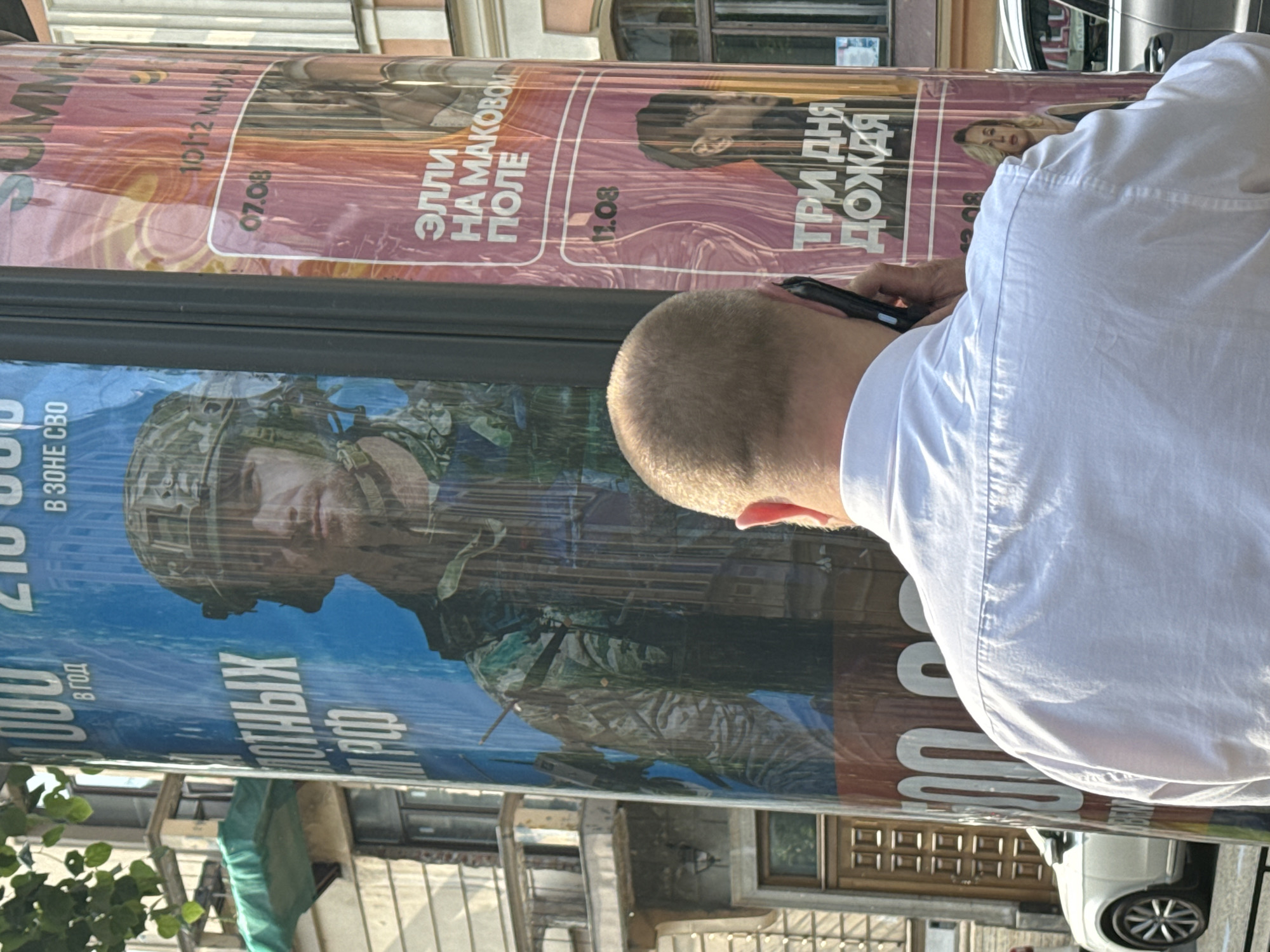 A man in a white shirt is on the phone, his back to the camera, with a public advertisement pillar to his right. The pillar displays two large posters. The poster on the left features a soldier in camouflage and helmet, with text in Russian that appears to be "В ОДНОЙ РОТЫ". The poster on the right has pink and yellow hues and shows text in Russian including "ЭЛЛИ НА МАКОВОМ ПОЛЕ" and "ТРИ ДНЯ ДОЖДЯ". The advertisements are on a street in Saint Petersburg, Russia, indicated by the architectural style of the building visible in the background, which features ornate detailing and a balcony. A white car is parked on the street to the left.