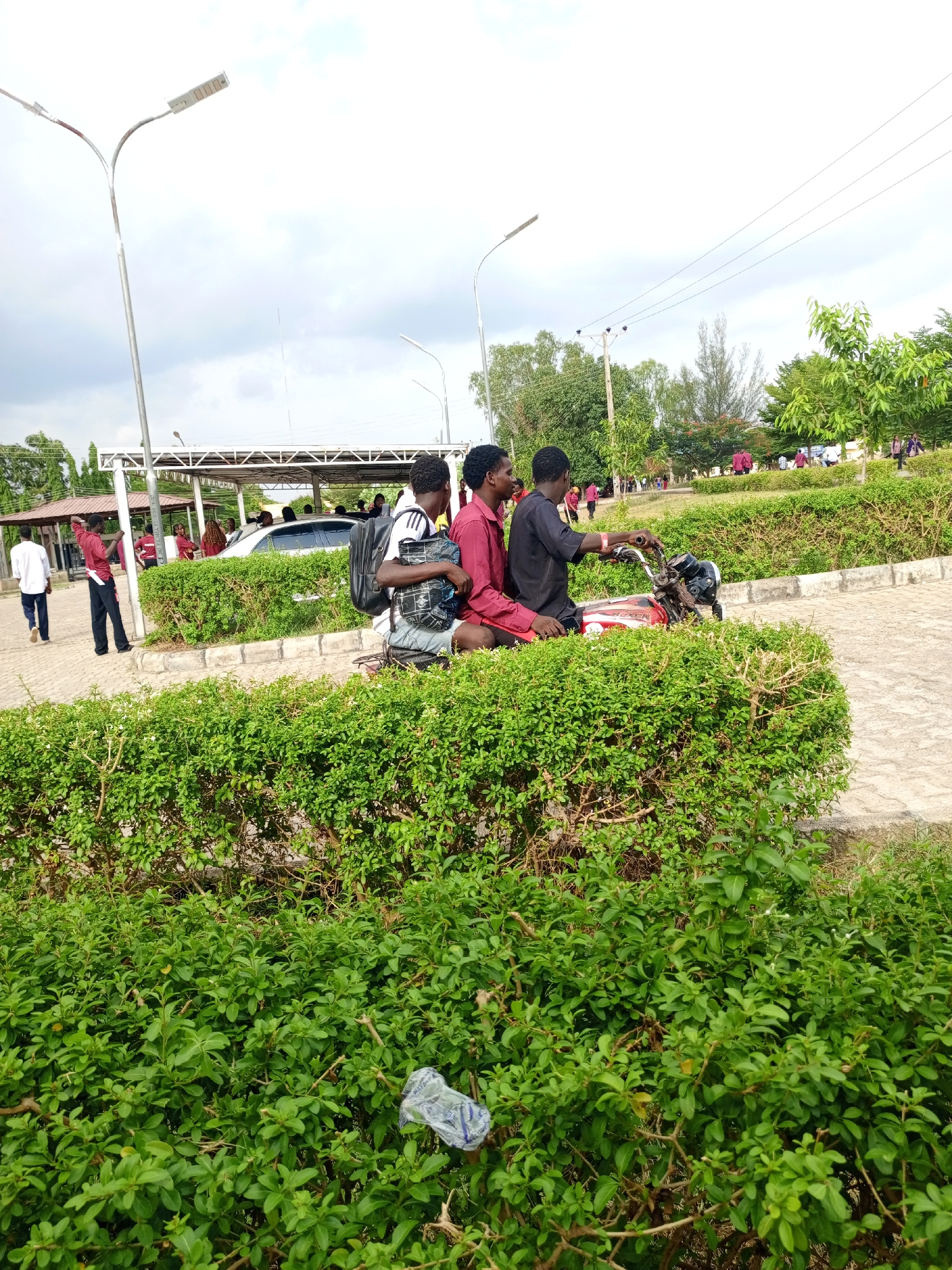 Three young men are on a red motorcycle in an outdoor setting, likely in Keffi, Nigeria. The motorcycle is partially obscured by a green bush. The man at the front is driving, wearing a dark shirt. The middle passenger wears a red shirt, and the rear passenger has a backpack. In the background are buildings, people walking, a covered parking area with a car, and trees. Streetlights are also visible. A discarded plastic bag is visible within the lower bush.