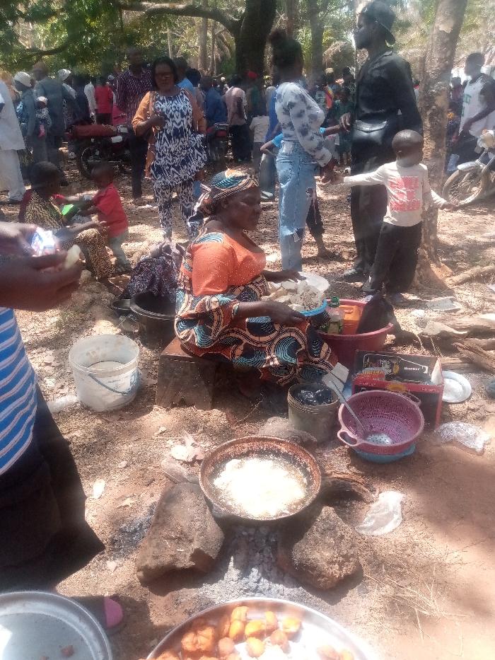 The scene depicts an outdoor gathering in Kagoma, Nigeria, characterized by food preparation and a crowd of people under a canopy of trees. In the mid-foreground, a woman, clad in an orange top, patterned skirt, and headwrap, is seated on a low dark block. She holds a white bucket filled with light-colored, diced food. Directly in front of her, a dark frying pan containing bubbling oil and cooking light-colored items rests on three large stones over an open fire with visible embers. Surrounding this cooking area are various containers including a white plastic bucket with liquid, a black pot with dark contents, a pink plastic colander, and a silver tray holding fried golden-brown food items (possibly plantains or yams) at the bottom of the frame.
Numerous individuals are present in the mid-ground and background. A person in a blue and white striped shirt is partially visible in the immediate foreground, handling a piece of light-colored food. Behind the seated woman, a woman in a black and white patterned dress with an orange jacket stands. Further back, a woman in a light patterned top and ripped blue jeans stands, while a man in a dark shirt and pants stands next to a small child in a white shirt. Two other young children are seated on the ground to the left. The background reveals a dense congregation of adults and children, with at least one motorcycle visible. The ground is unpaved earth, consistent with an outdoor setting under trees providing shade, and the lighting indicates daytime.