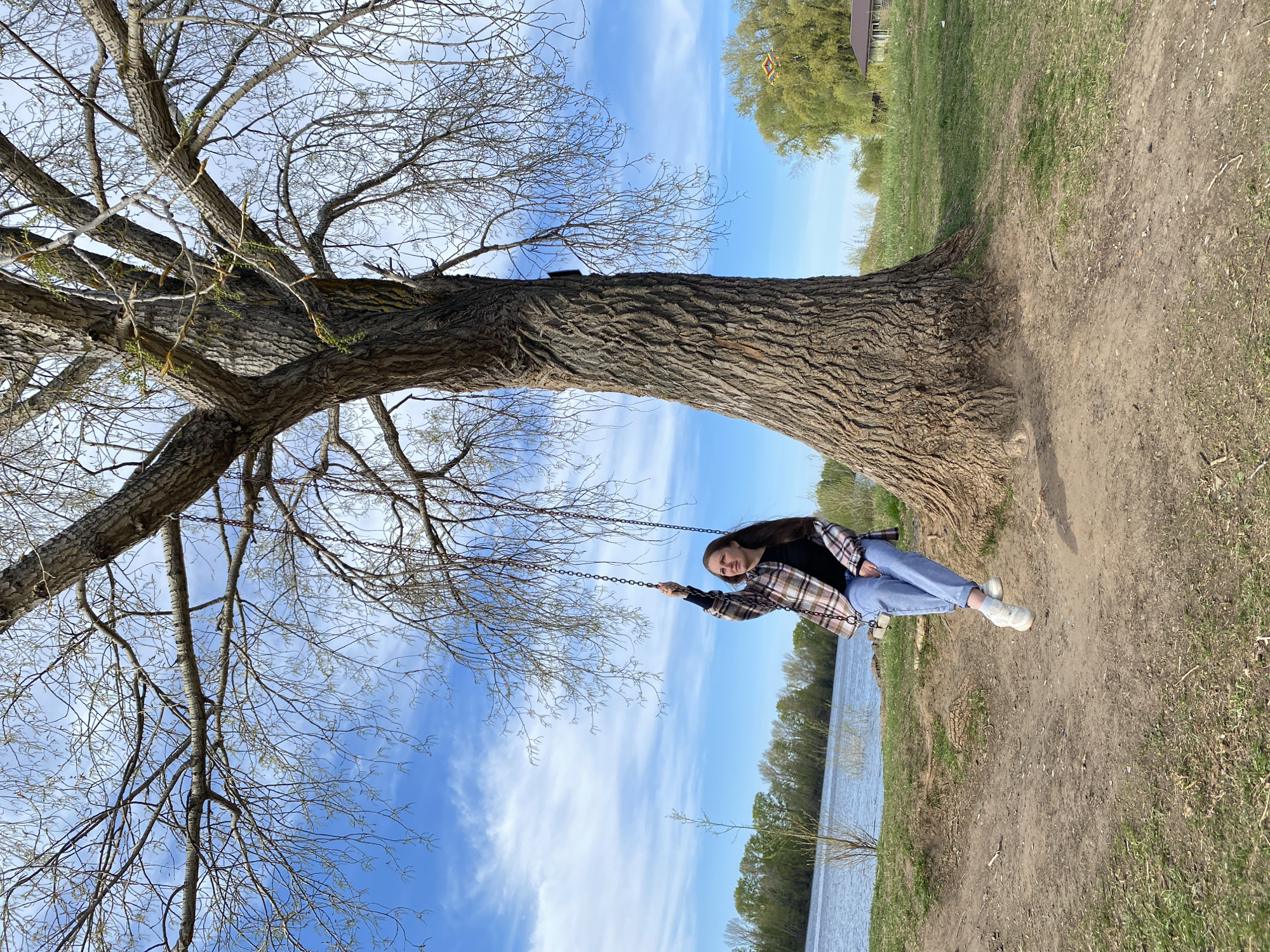 A woman is on a swing suspended from a large tree in an outdoor setting. She is wearing blue jeans, white sneakers, a black top, and a plaid shirt. The tree has a thick, textured trunk and bare branches reaching towards a blue sky with wispy clouds. In the background, there is a body of water and some greenery. The ground is dirt and grass. The location is outdoors in Chaikovsky, Russia.