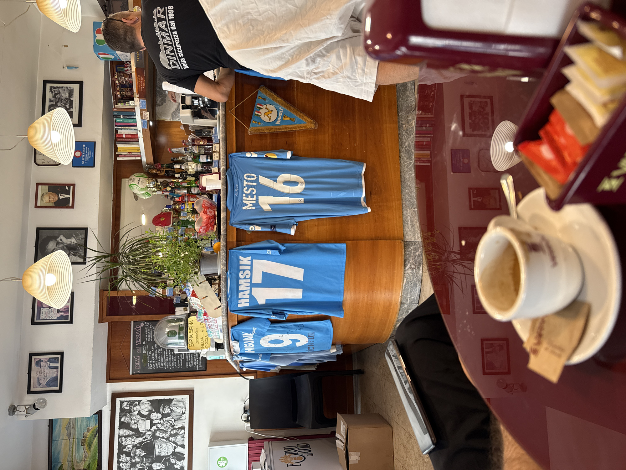 The image depicts the interior of a bar, likely in Naples, Italy, given the presence of Napoli football jerseys and a Napoli pennant. The bar features a wooden counter with various bottles of alcohol, a coffee machine, and a shelf stocked with books and decorative items. Several framed photographs and posters adorn the walls, including portraits and a landscape. In the foreground, a table with a red reflective surface holds a white espresso cup and saucer, along with a small box containing sugar packets and what appear to be pastries. A person wearing a black t-shirt with "DINMAR" printed on it is visible behind the counter, facing away from the camera. Three light blue Napoli football jerseys with player names (HIGUAIN, HAMSIK, MESTO) and numbers are hanging on the counter. The overall ambiance suggests a casual, perhaps sports-themed, establishment.