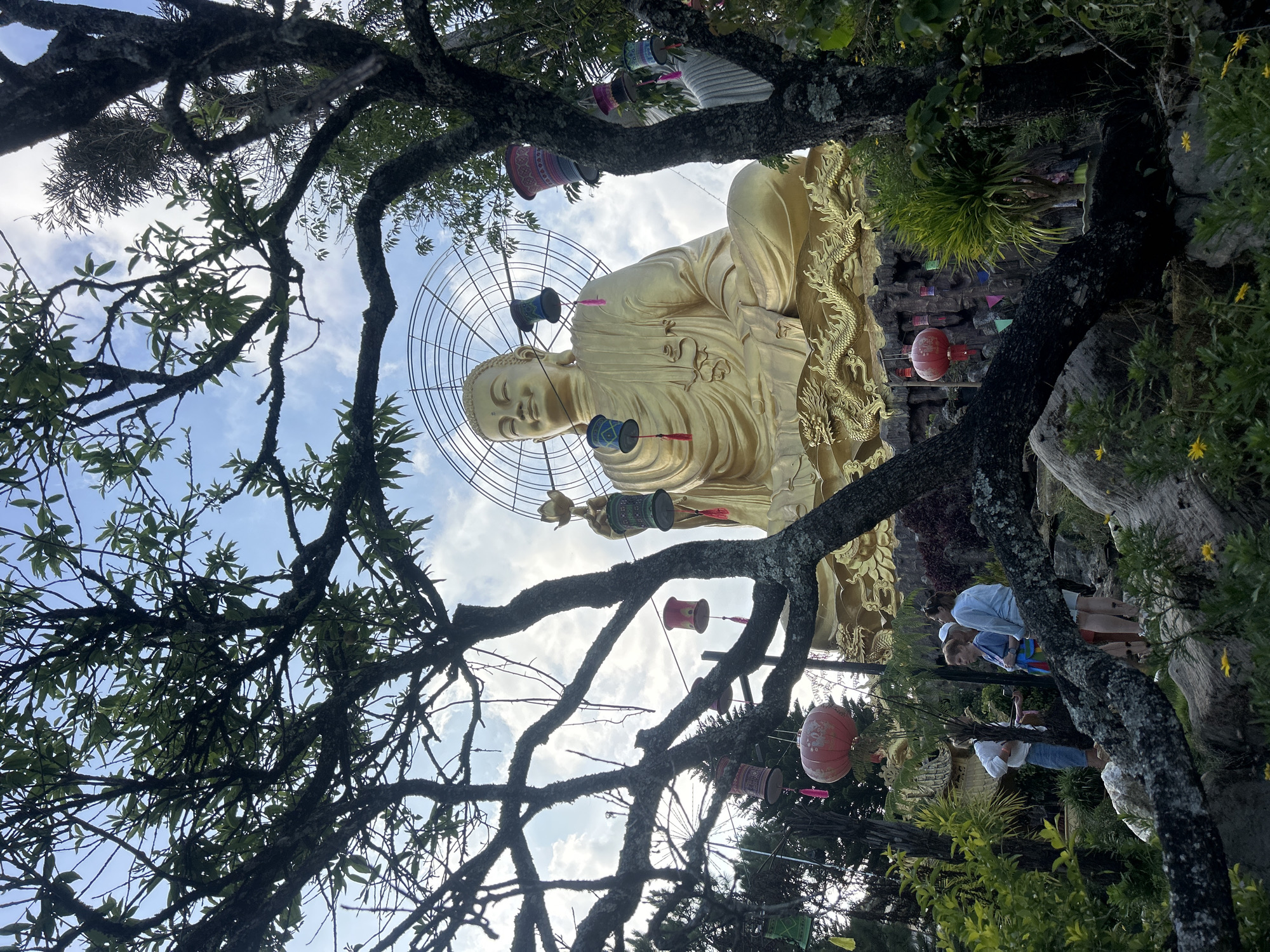 The photograph, taken in Lâm Viên - Đà Lạt, Vietnam, presents a low-angle view of a golden Buddha statue framed by the branches of a tree. The statue is positioned in the center of the frame. Several decorative ornaments, including lanterns, hang among the tree branches. To the right of the frame, partially visible, are three individuals dressed in casual attire. The background includes hints of other plants and structures, and patches of sky are visible through the tree's canopy.
