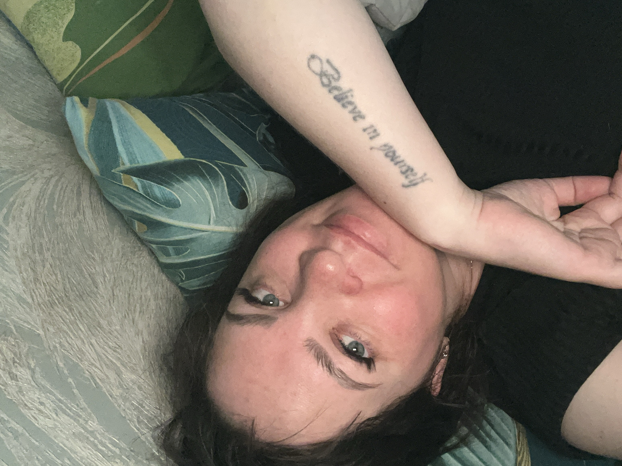 A fair-skinned woman lies on a textured, light-colored surface with two pillows: one patterned with green, brown, and yellow stripes, and another featuring a blue and green leaf design. She wears a black garment and has dark hair. Her left arm is raised, revealing a tattoo that reads "Believe in yourself" in cursive script. She has blue eyes and wears eyeliner. The image does not contain location information regarding the city of Noviy, Russia.