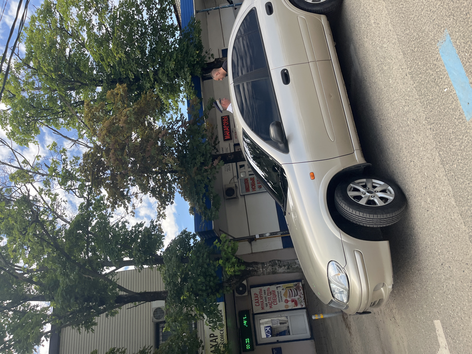 The image, presented sideways but depicting an upright scene, captures a bright daytime moment in Krasnodar, Russia. A light-colored (beige or light gold) sedan is parked on asphalt pavement next to a building. The sky is bright blue with scattered white clouds, and the area is densely populated with lush green trees, some showing signs of brown leaves, bordering the street and partially obscuring the building.
Two men are visible near the passenger side of the car. One man, bald and wearing a dark shirt, stands close to the vehicle. The second man, wearing a white hat or cap and a white shirt, stands slightly further back, seemingly at the entrance of the building. Their interaction is not entirely clear but they appear to be near a shop.
The building is a commercial establishment, likely a small grocery or convenience store. Multiple signs in Russian are visible. A large banner advertises "САХАР, МУКА, КРУПЫ, ЧАЙ, КОФЕ, СОКИ, МАСЛО, СОЛЬ, СПЕЦИИ, КОНСЕРВАЦИЯ, СУХОФРУКТЫ" (Sugar, Flour, Groats, Tea, Coffee, Juices, Oil, Salt, Spices, Canned Goods, Dried Fruits). Other signs feature "МАКАРОНЫ" (Macaroni) and "НОВЫЕ" (New). A digital display shows the temperature as "24°C" and the time as "09:31". Air conditioning units are mounted on the building's facade. The overall scene suggests a routine morning or midday in an urban or suburban setting.