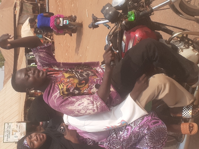 The scene depicts an outdoor, dusty street in Gwadanye, Nigeria, under clear daylight, featuring multiple individuals and motorcycles. In the foreground, three people are positioned on a red motorcycle, which appears stationary or moving slowly. The driver, a dark-complexioned male wearing a multi-colored purple graphic t-shirt and dark pants, has his left arm raised with an open hand, mouth open as if speaking or shouting, while his right hand rests on the handlebar. Directly behind him, a passenger in a light-colored long-sleeve top and light pants sits, with a hand visible on the driver's back. Behind this passenger, a dark-complexioned female wearing a black head covering and a patterned purple dress holds a smartphone in her left hand and looks towards the driver. The feet of the passengers are visible; the front passenger wears checkered slides, and the rear passenger wears brown sandals.
In the midground, an unpaved, reddish-brown dirt road extends into the distance, with another motorcycle moving away from the viewer. This second motorcycle carries two individuals, with a female passenger in a blue t-shirt and pink pants visible from the back. The background features several single-story structures with corrugated metal roofs, indicative of market stalls or residences. A partially obscured white sign with black text reading "WELCOME TO EVANGEL KUB PRIAO SECONDARY SCHOOL" is visible on the left side of the background. A green translucent object is attached to the front of the foreground motorcycle.