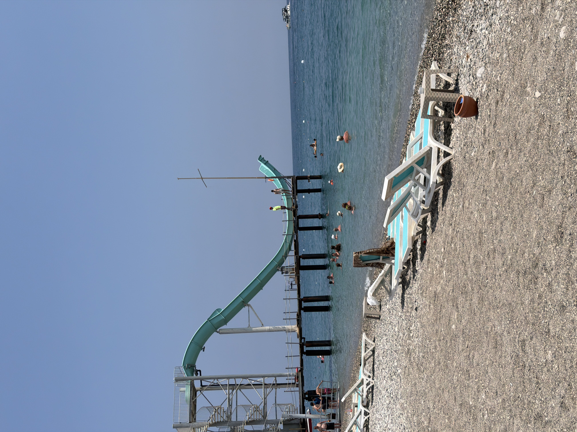 The image depicts a bustling beach scene in Kemer, Turkey, likely at a resort. A large, turquoise water slide dominates the left side of the frame, its structure extending over the water on a wooden pier. Several people are visible at the top of the slide, preparing to descend, while others are already in the water, swimming and enjoying themselves. The beach itself is covered in small pebbles, with rows of empty lounge chairs laid out for sunbathers. The turquoise waters of the Mediterranean Sea stretch out to the horizon, with a small boat visible in the distance. The sky is a clear, vibrant blue.