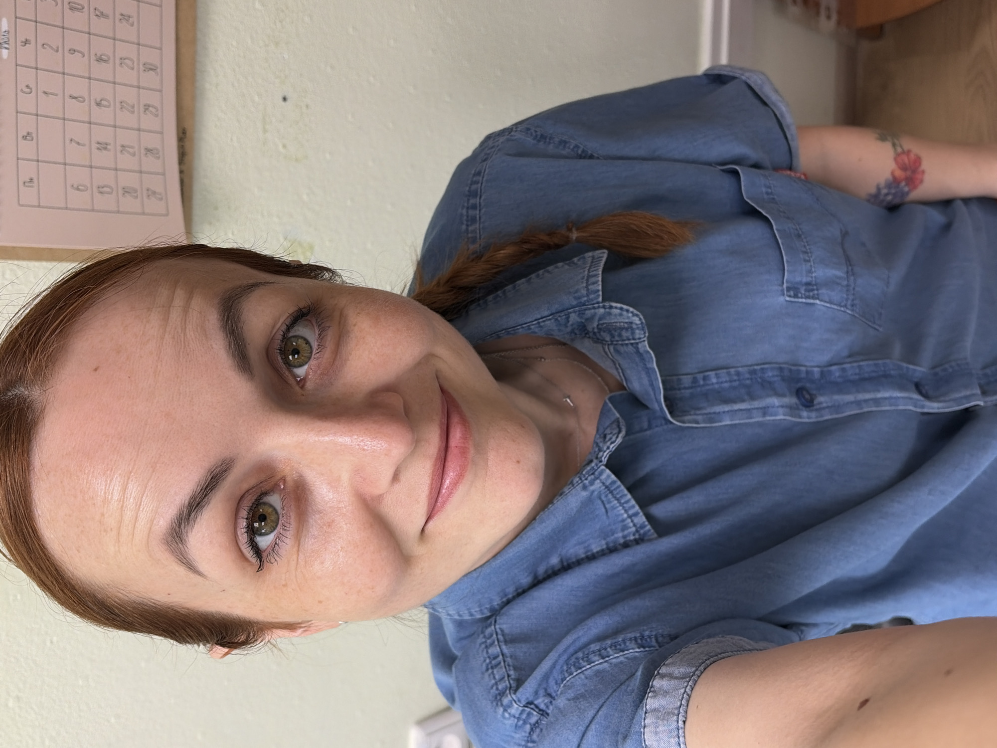 A woman with reddish-brown hair, a fair complexion, and light green eyes is looking at the camera and smiling. She is wearing a blue denim shirt. Her hair is pulled back and braided to her right. She has a few freckles on her face. There is a calendar on the wall behind her, which appears to be a monthly calendar with some writing and numbers. The wall behind her is textured and appears to be painted a light, neutral color. A small portion of a white electrical outlet is visible on the left side of the image. There is a floral tattoo on her left arm. The scene appears to be indoors, possibly in an office or residential setting. The location is Chaikovsky, Russia.