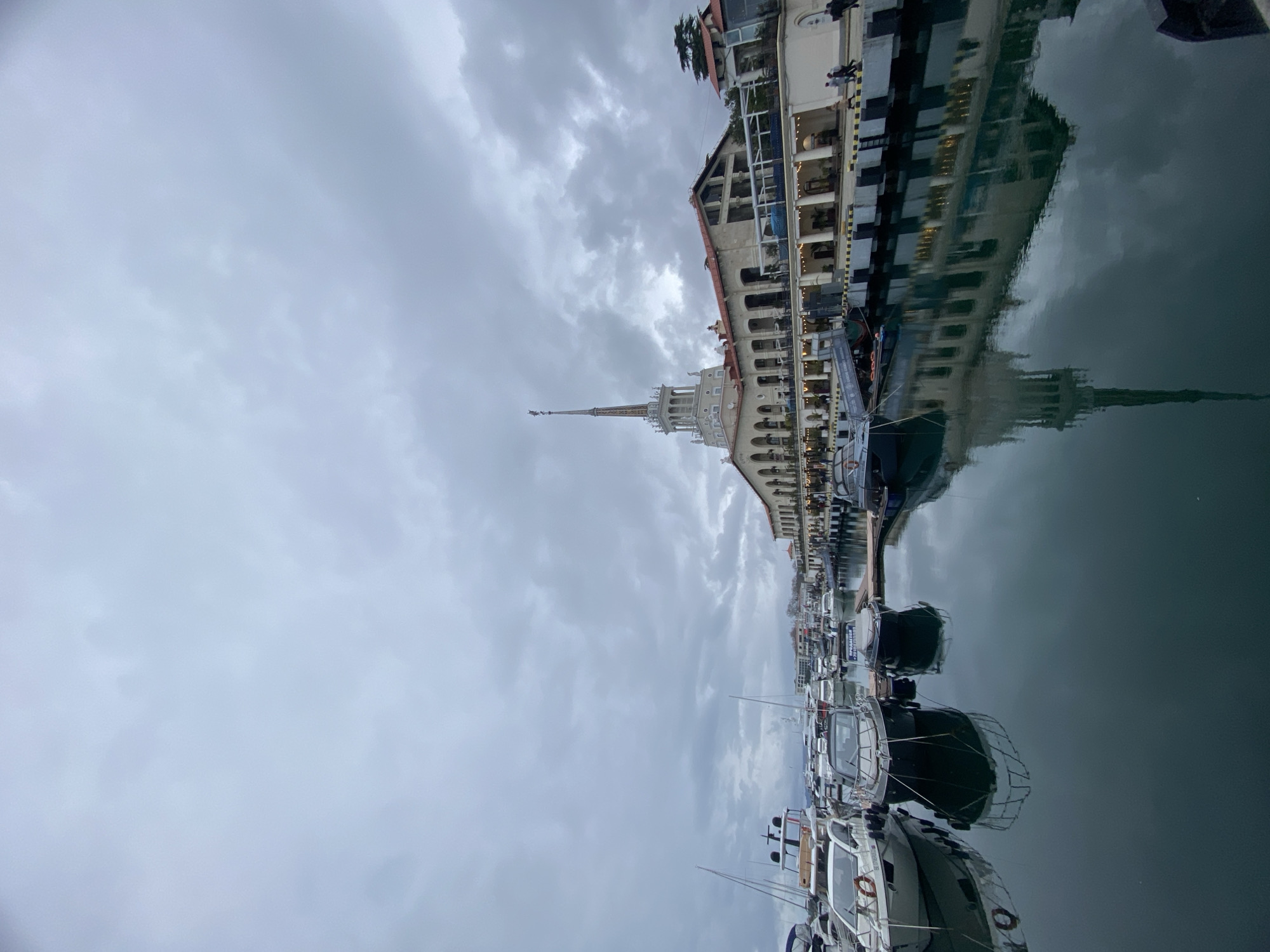The image depicts a marina in Sochi, Russia, under a cloudy sky. The water reflects the architecture, creating a mirrored effect. Rows of docked yachts and boats are visible along the bottom of the frame. The Sochi Marine Station, characterized by its tall spire and white facade, stands prominently, its reflection visible in the water. The scene is devoid of people.