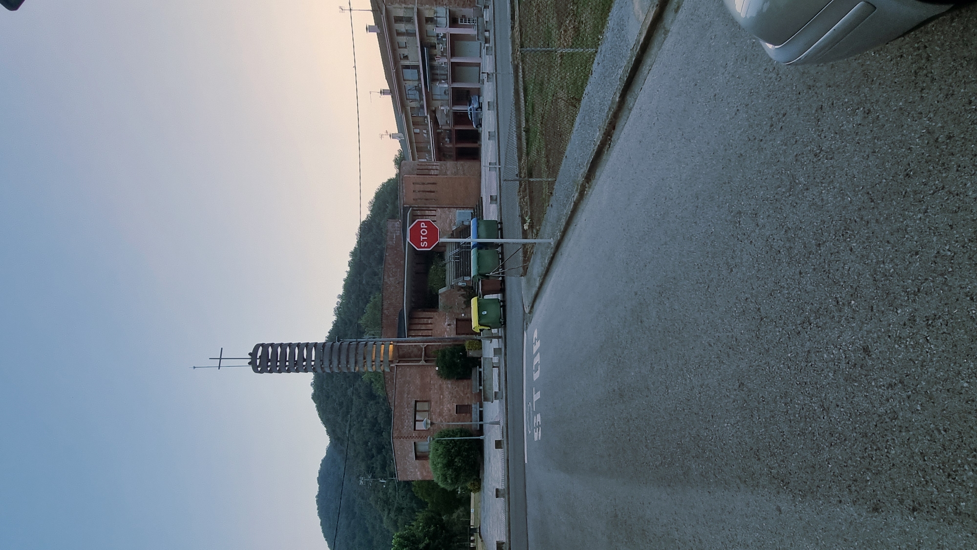 The image depicts an outdoor scene in La Canya, Spain, likely during twilight. In the foreground, a paved road curves towards the right, with a silver car partially visible on the right edge. White painted text on the road reads "STOP". In the middle ground, a red octagonal stop sign stands on a metal pole. Behind the stop sign are several green and brown trash receptacles. A low concrete barrier with bollards runs along the edge of the paved area. Further back, a brick building with a distinctive, modern tower topped with a cross is visible. The tower features a spiraling metal structure. Adjacent to this building, another multi-story residential building with balconies can be seen. The background consists of a lush, tree-covered hillside under a pale sky. The overall impression is one of a quiet, residential or community area.