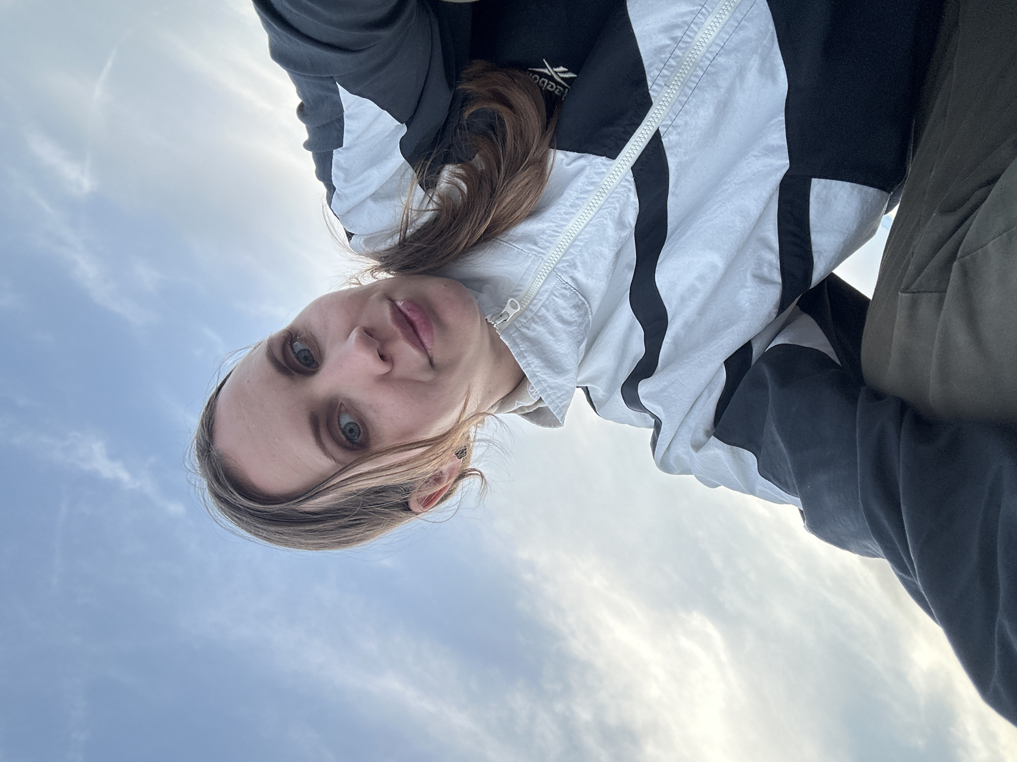 An upward-facing selfie shows a woman with fair skin, light brown hair, and light blue eyes. She wears a Reebok jacket with black and white panels, partially zipped, and a dark garment underneath. She is looking at the camera. The background consists of a light blue sky with wispy clouds, suggesting an outdoor setting. Due to the lack of landmarks, pinpointing a specific location in Fethiye, Turkey, is impossible.