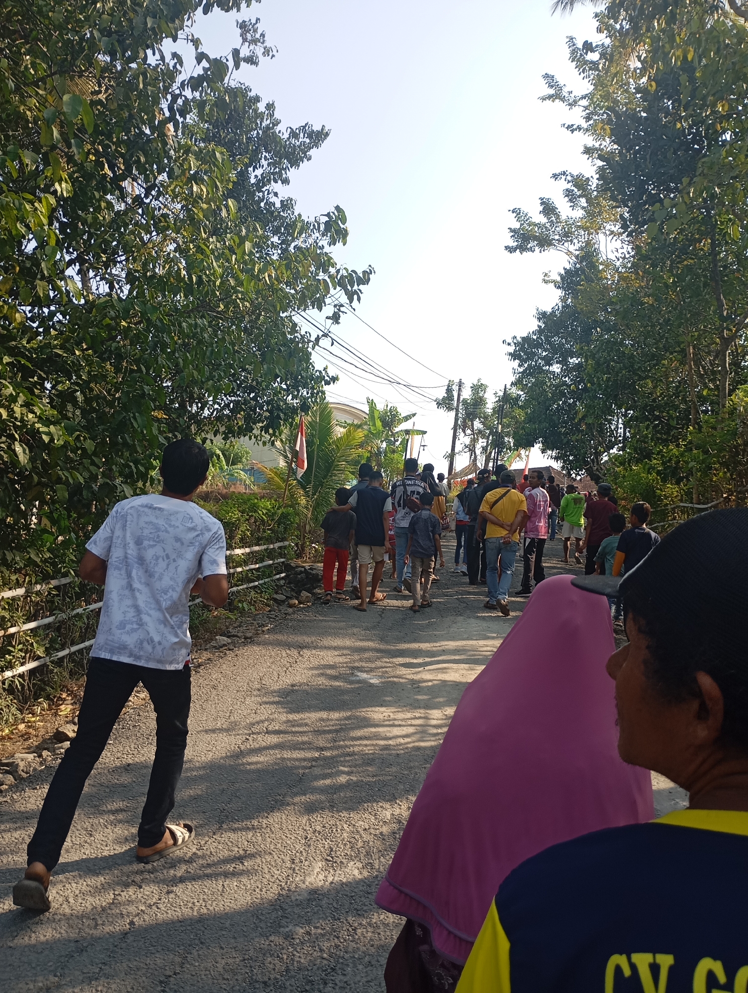 The image captures a street scene in Ketepung, Indonesia, on a clear day. A group of people are walking along a paved road lined with lush green trees and vegetation on both sides. In the foreground, a person in a white t-shirt with a patterned design is walking away from the camera, their back to the viewer. To the right, a person wearing a pink headscarf is also visible, with another person in a blue and yellow shirt partially in frame. The group of people further down the road are walking in the same direction. Several individuals are wearing casual clothing, including t-shirts, shorts, and pants. One person is wearing a yellow t-shirt, and another a pink t-shirt. A flag, appearing to be the Indonesian flag, is planted on the left side of the road amidst tropical foliage. Electrical wires are strung overhead, suggesting the presence of infrastructure. The overall atmosphere suggests a casual gathering or movement through a local neighborhood.
