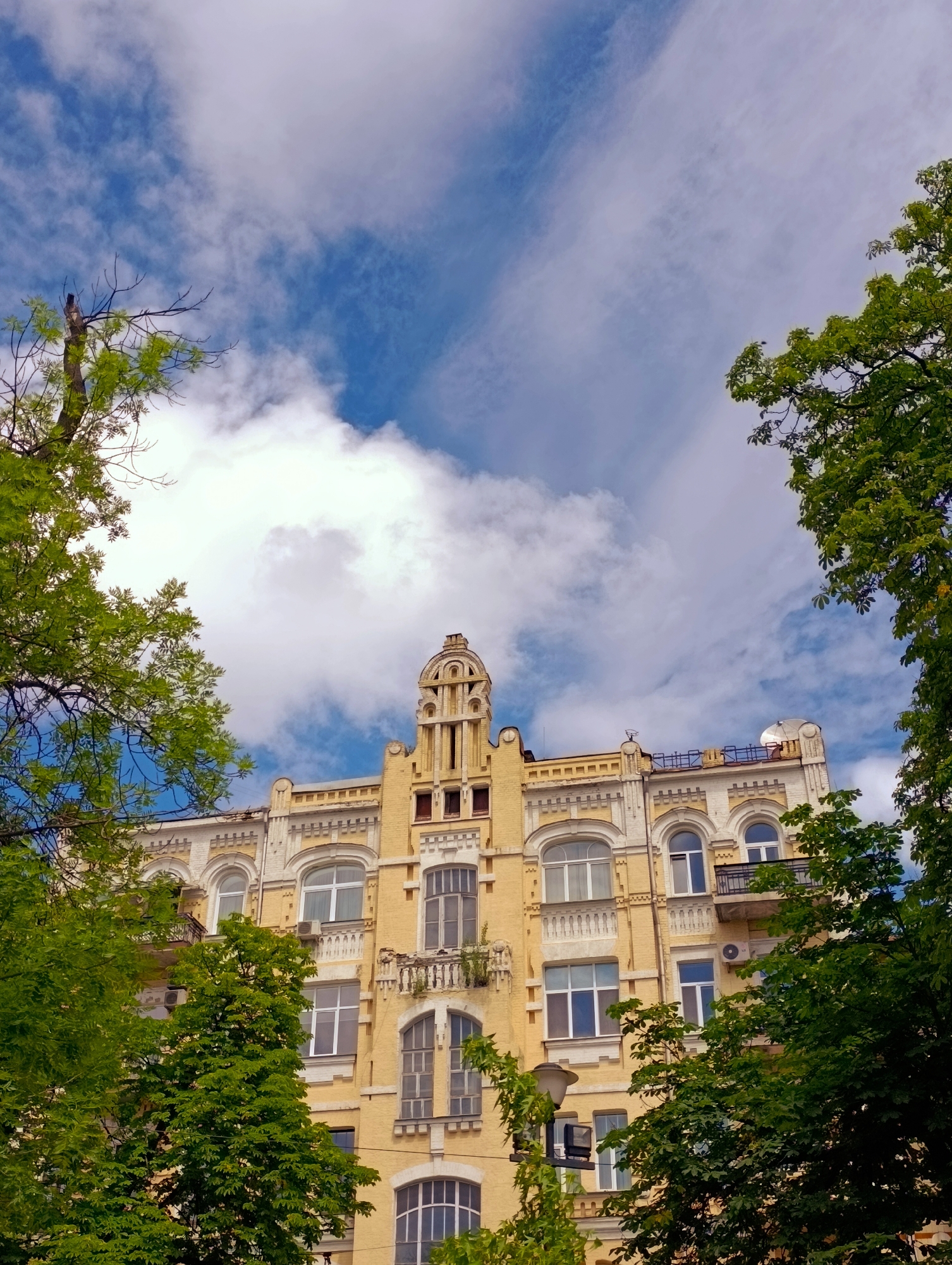 A low-angle shot captures the facade of a yellow, multi-story apartment building in Kyiv, Ukraine, framed by lush green trees and a sky filled with white, wispy clouds against a bright blue backdrop. The building features an ornate, Art Nouveau-inspired design with decorative elements around the arched windows, balconies, and a prominent tower-like structure on its upper level. The windows are a mix of old and new, with some appearing to be original wooden frames and others modern replacements. Air conditioning units are visible on the facade. In the foreground, the branches and leaves of several trees obscure parts of the building, suggesting a verdant urban environment. A lamppost with two lights stands to the left of the building, partially covered by tree branches. The overall impression is one of an older, architecturally interesting building surrounded by nature in a city setting.