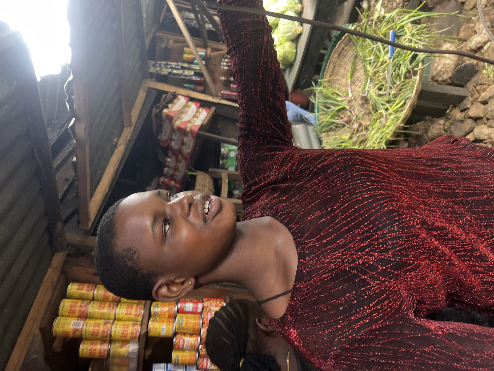 A young Black woman, wearing a shimmering red long-sleeved top, is in a market stall in Nkpor, Nigeria. She is looking to her right and speaking, with her mouth open slightly, revealing teeth. Her hair is short and dark. Behind her, to the left, is another Black person, also with dark hair, facing away from the camera.
The market stall is rustic, with a corrugated metal roof and wooden shelving. On the shelves are rows of yellow cans, some with red labels that appear to say "BAKED BEANS." Further back, there are boxes of products with red and white packaging. In the foreground and to the right, a large woven basket is filled with green onions and what appear to be small shallots. Cabbages are visible to the right of the basket. The ground appears to be rocky and uneven. The lighting suggests it is daytime, with sunlight filtering through gaps in the roof.