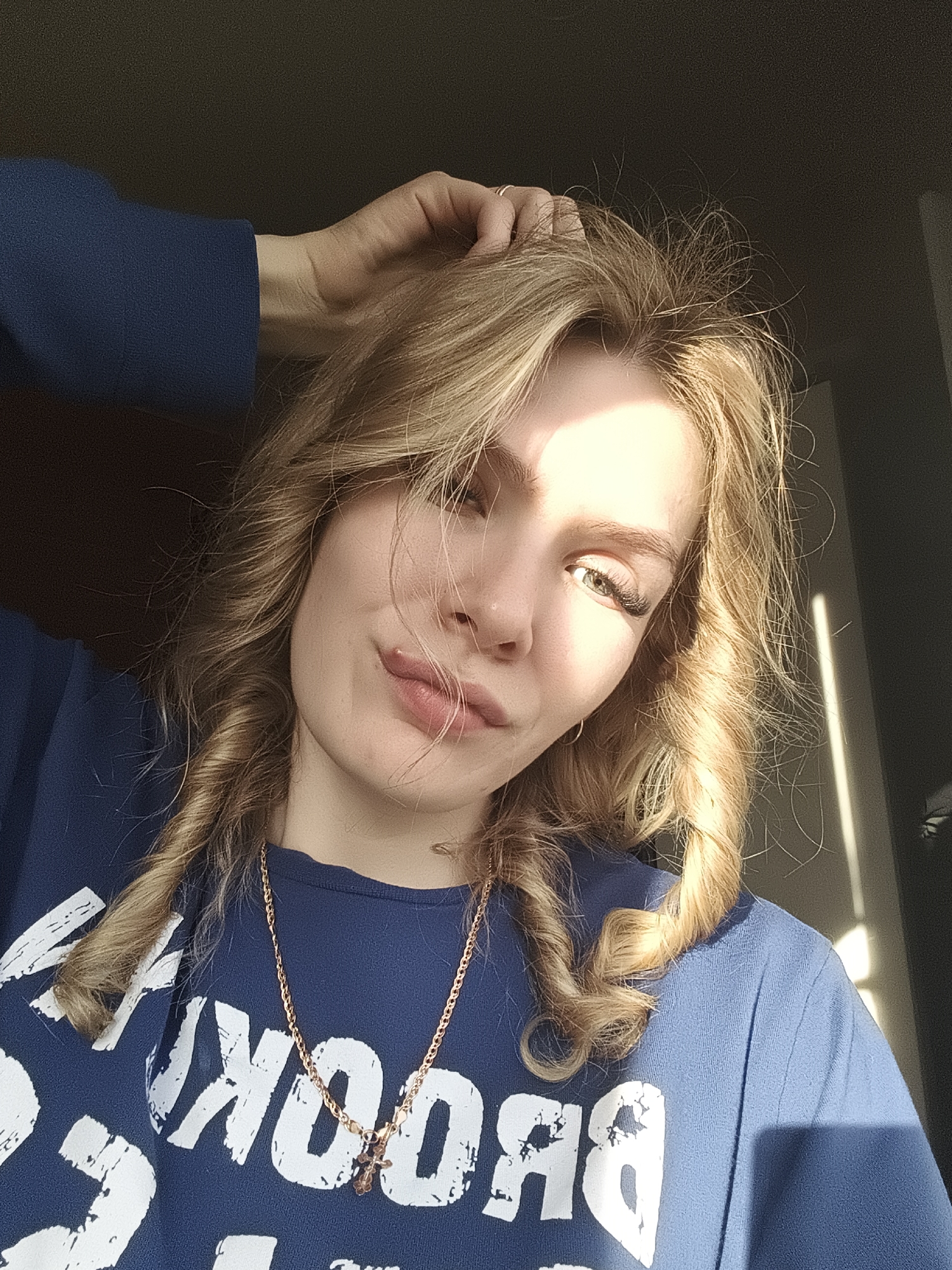 A woman with blonde, shoulder-length, curly hair poses for a selfie. She is wearing a blue long-sleeved shirt with white lettering, and a gold-colored necklace with a pendant. Her left hand is touching her hair near the crown of her head. Her gaze is directed towards the camera. Sunlight illuminates the right side of her face and hair. The background is a dark room with a door or window on the right, where sunlight is visible. The location appears to be indoors, but there is no clear evidence to definitively place it in the city of Русский Вожой, Russia.