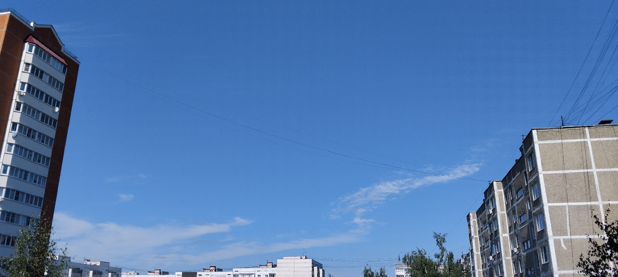 The image shows a view of apartment buildings against a clear blue sky with thin clouds. On the left is a tall, multi-story apartment building with a brick exterior and white window frames. To the right, another apartment building with a gray concrete facade and white grid patterns is visible. Power lines crisscross the sky. Trees are visible at the bottom of the image. The scene appears to be a residential area in a city.