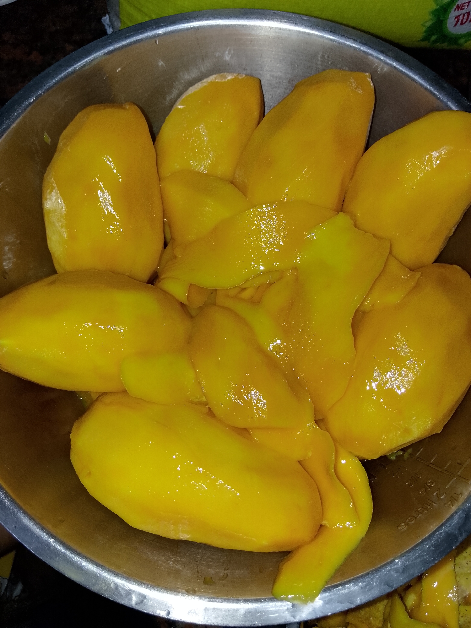 In a stainless steel bowl, a collection of peeled mangoes are arranged. The mangoes are a vibrant yellow, with some pieces showing a slightly lighter hue in certain areas. The flesh of the mangoes appears smooth and moist, with a natural sheen reflecting the light. The bowl itself is circular with a metallic finish and a visible rim. The interior of the bowl also displays measuring lines and text in both imperial and metric units, suggesting it might be a mixing or measuring bowl. The background is out of focus, with a hint of a green object and some dark areas. The image focuses on the mangoes and the bowl, creating a close-up view of the fruit. The scene appears to be indoors.
