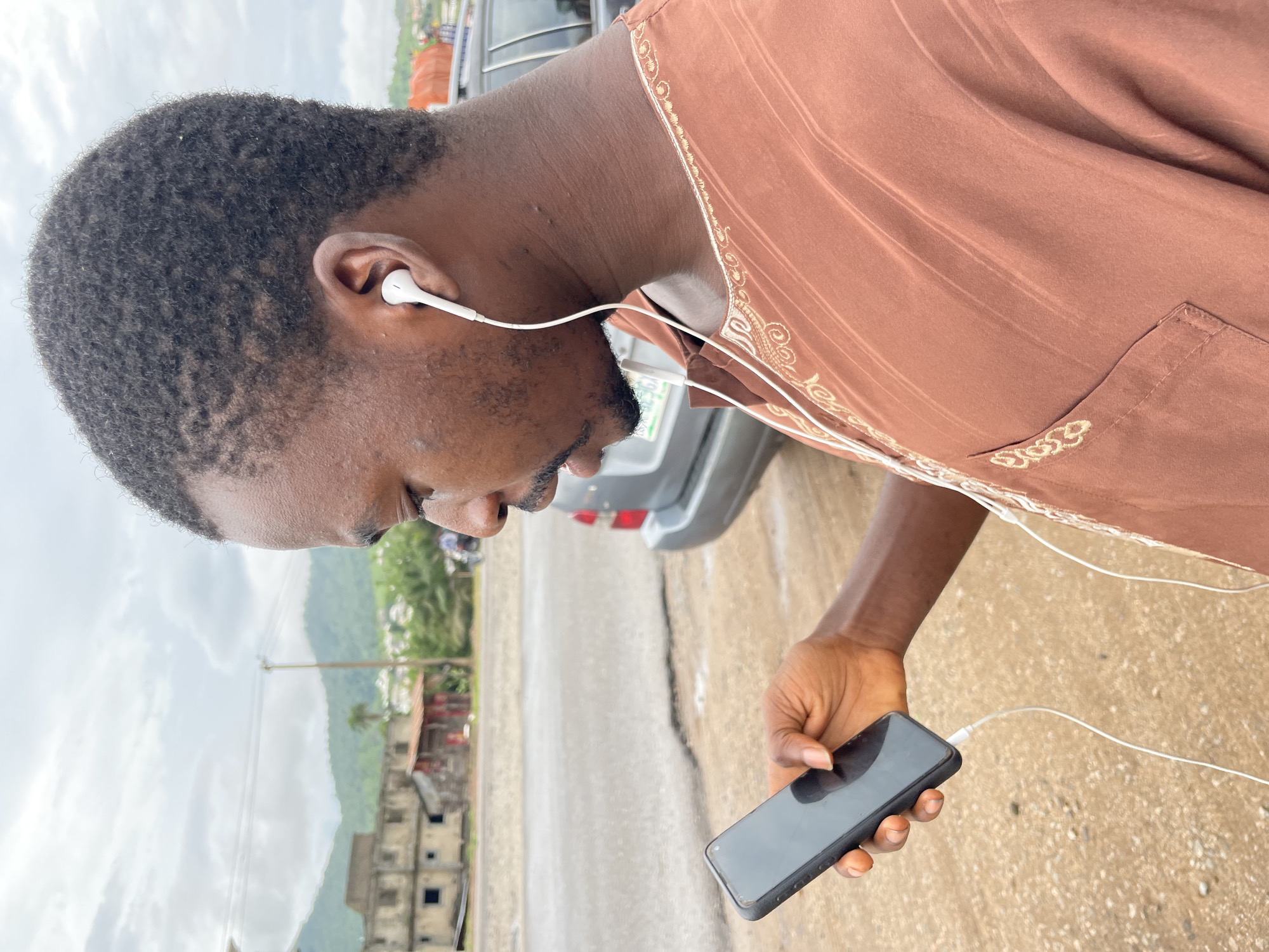 The media file shows a close-up, side-profile of a person wearing white earbuds and holding a black smartphone. The person is wearing a brown garment with decorative gold trim and a pocket. The background suggests an outdoor setting in Lokoja, Nigeria, with a road, buildings under construction, and lush green hills under a cloudy sky. The time of day appears to be daytime, with overcast weather conditions. The person's focus is on the phone, suggesting they are engaged in listening to audio or using an application. No visible text is present in the image.