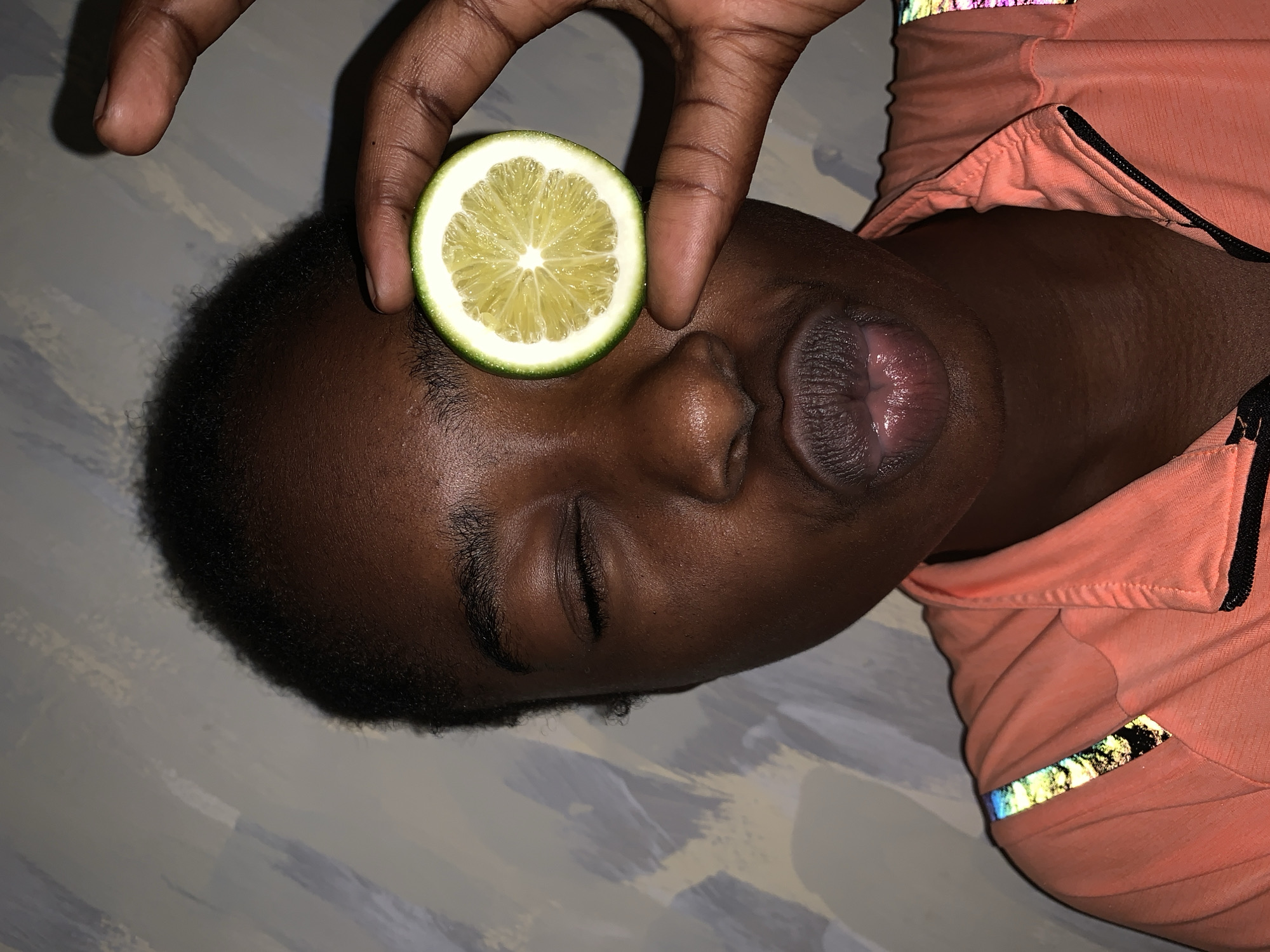 A dark-skinned individual is lying on a surface, with a slice of lemon placed on their forehead near the eyebrow. A hand holds the lemon in place. The person's eyes are closed and their mouth is slightly open. They are wearing an orange garment with black trim and reflective details. The background appears to be a textured surface, possibly a painted floor or wall. The image is taken from a top-down perspective. There is no information in the image to suggest a location in the city of Rafin Par, Nigeria.