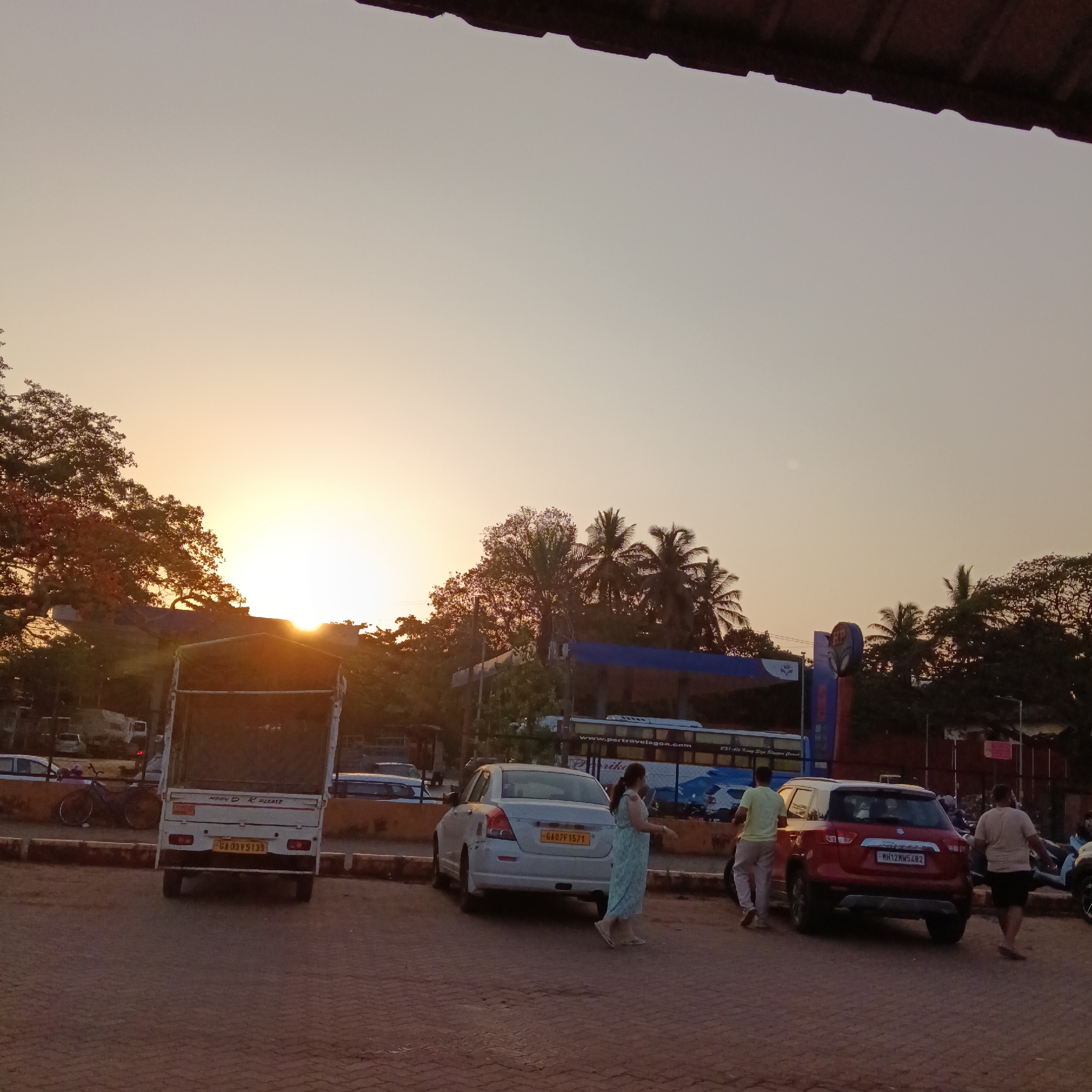 The image captures a street scene at dusk in Mapusa, India. The sky is illuminated by the setting sun, creating a warm, golden hue. In the foreground, there is a paved area with several parked vehicles, including a white utility vehicle, a white sedan, and a red SUV. Three people are visible in the image: a woman in a light-colored dress walking towards the left, a man in a light-colored shirt and pants walking towards the SUV, and another man in a light-colored shirt and dark shorts walking away from the SUV. In the background, there is a petrol station with a bus parked in front of it, as well as trees.