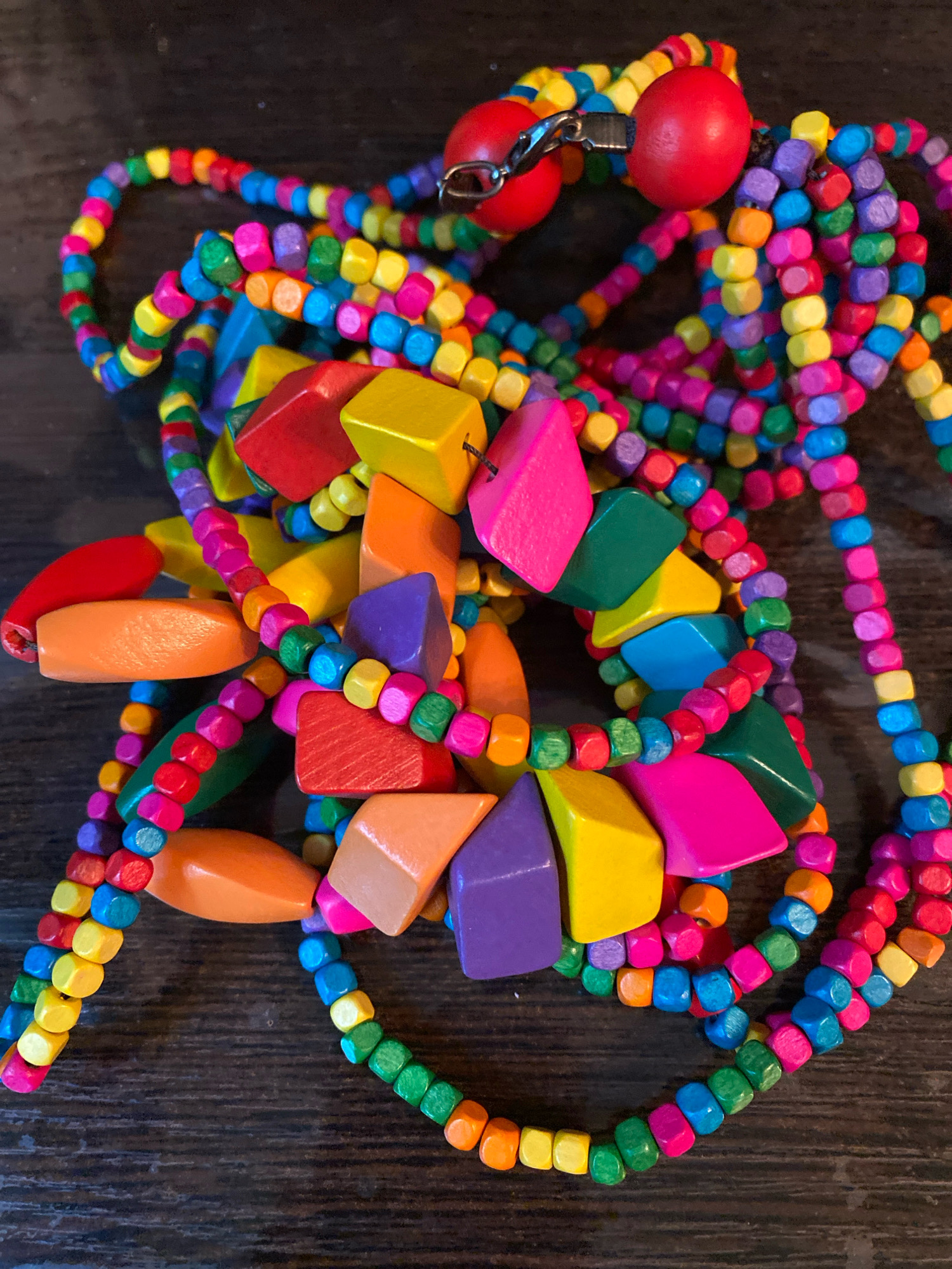 The image displays a close-up, overhead view of a tangled assortment of colorful, wooden beads and shaped wooden pieces, likely part of jewelry or craft supplies. The beads are small and cubic, in various shades of green, blue, purple, yellow, orange, and red. Interspersed are larger, flattened, geometric wooden pieces in bright pink, purple, yellow, orange, and red. Two smooth, red, spherical beads are visible at the top center, connected by a silver clasp. The objects are arranged on a dark, textured surface, possibly wood. The background is dark and out of focus. There are no people or animals present, and no discernible location in the city of Ivanteyevka, Russia. The scene is static and appears to be a still life arrangement of craft materials.