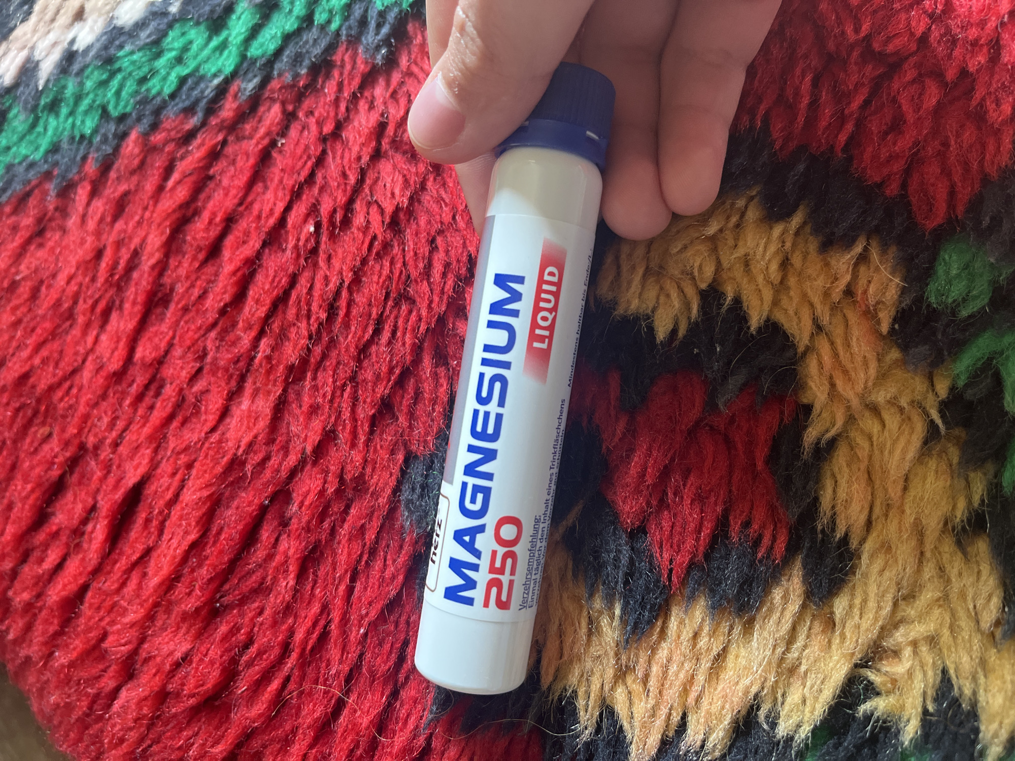 A human hand holds a small, white plastic bottle labeled "MAGNESIUM 250 LIQUID" against a patterned, multi-colored rug. The rug displays red, black, yellow, and green colors. The bottle has a blue cap. Text in German is printed on the bottle. The setting is possibly a household. There is no specific information given about Lozarevo, Bulgaria.
