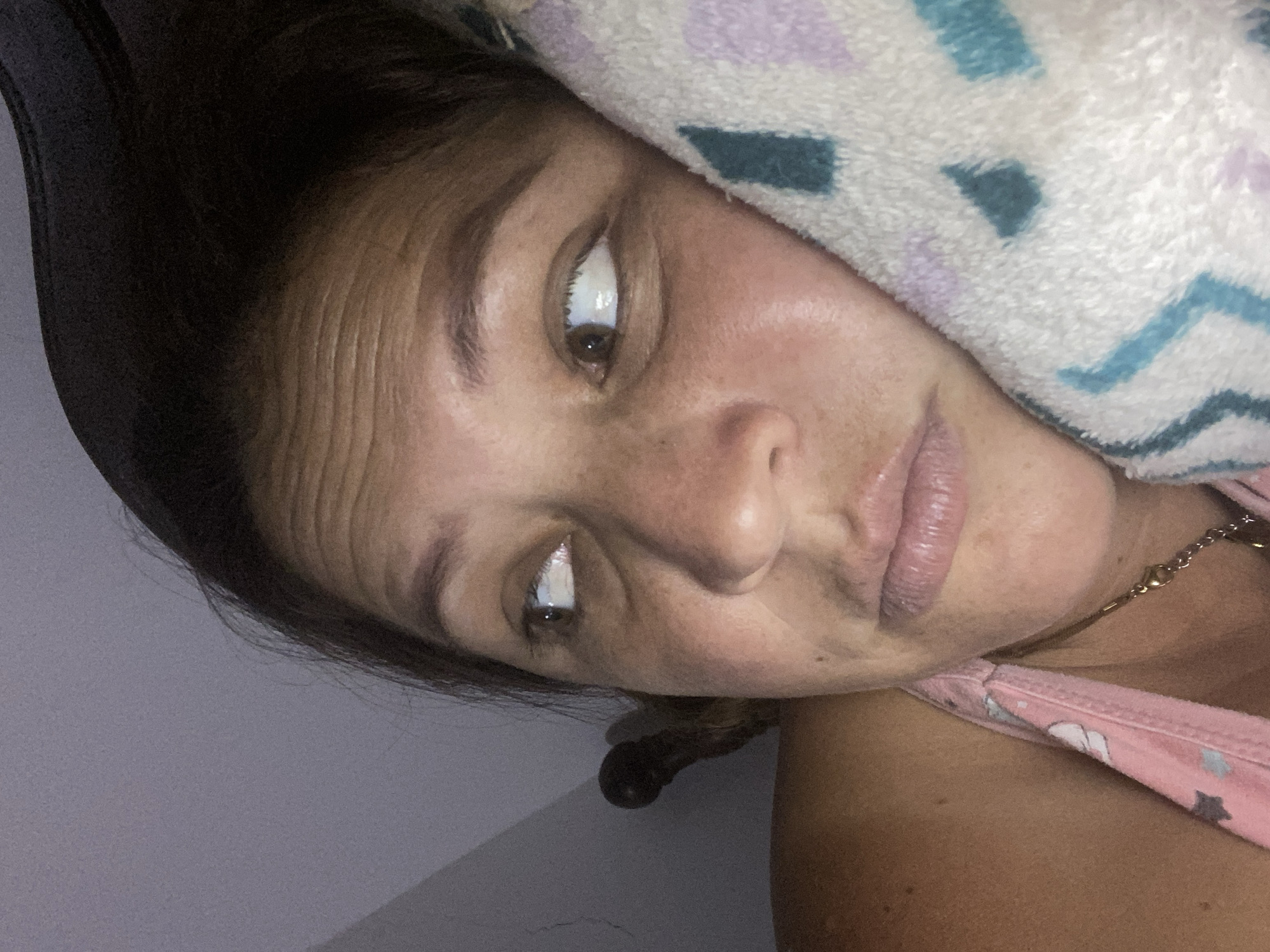 A close-up, slightly tilted portrait shows a person's face and upper body. The person is lying down, with their head resting on a patterned towel that has blue, purple, and teal geometric shapes. Their hair, dark brown, is visible on the left side of the frame, partially covered by the towel. The person's eyes are open and directed upwards, with one eye clearly visible and the other partially obscured by the towel. Their skin appears to have freckles and slight wrinkles on the forehead. The person is wearing a pink tank top with a star pattern. A thin gold chain necklace is visible around their neck. The background is a plain, muted gray wall. The lighting suggests it could be indoor lighting, possibly during the day or evening. The overall mood appears casual and relaxed. There is no visible text in the image.