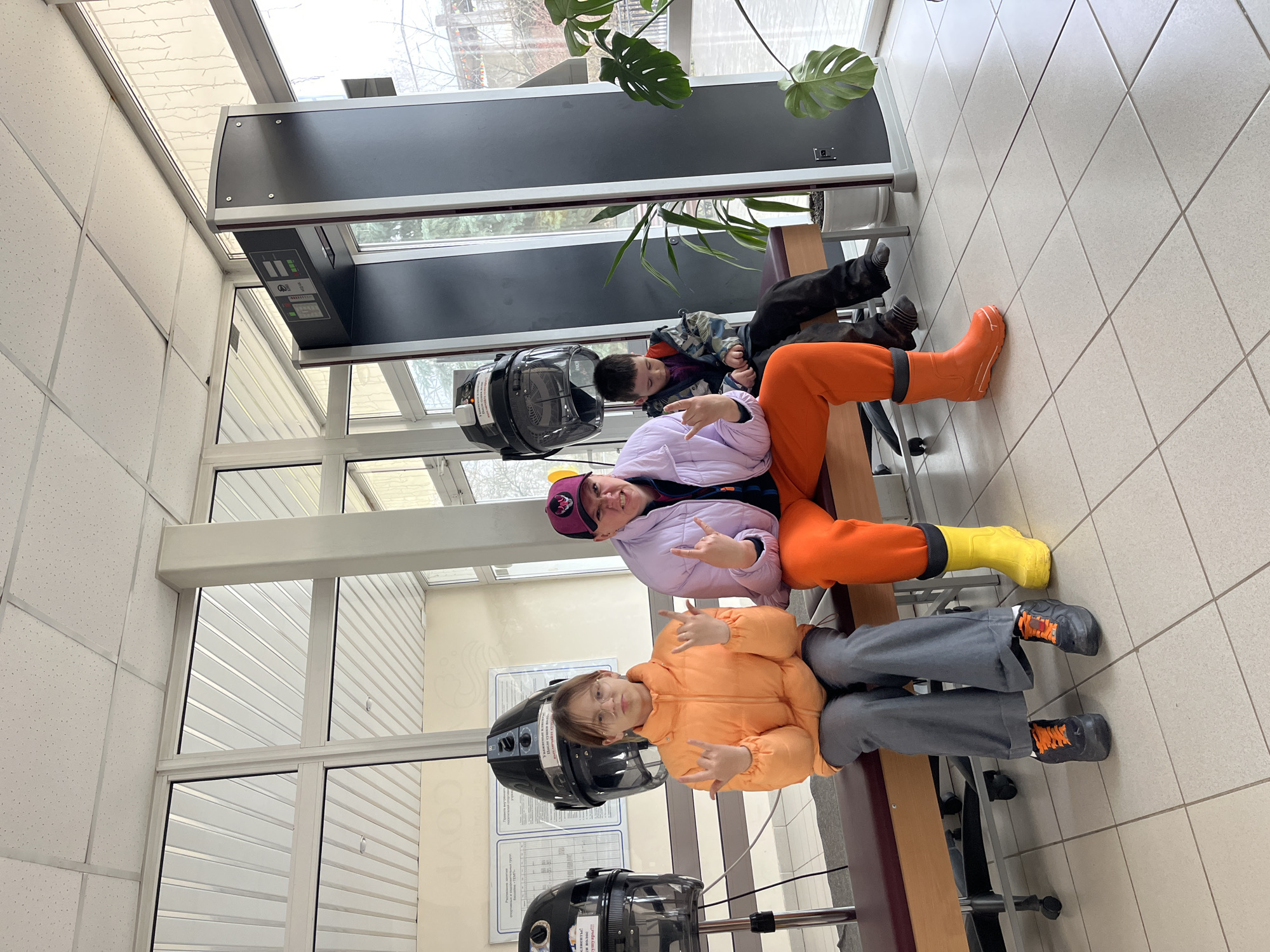 This image captures three children or young people seated on a long, brown bench in a brightly lit indoor space, likely a vocational training room, salon, or community center in Chaikovsky, Russia. The setting features large windows that reveal bare trees and an overcast sky outside, suggesting daytime during a cooler season.
In the foreground, a child with glasses wears an orange puffer jacket and gray pants with black shoes, making a "rock on" hand gesture. Beside them is a black stand-alone hair dryer with a clear plastic hood. Another child is seated next to them, wearing a light purple puffer jacket, bright orange pants, and yellow and orange rubber boots, also making a "rock on" gesture while sporting a purple and yellow cap. A second black hair dryer is positioned near this child. Further back and to the right, a third child in a camouflage jacket and dark pants is visible, appearing to rest or look down.
The room has a tiled floor and an acoustic tile ceiling. A large, dark gray rectangular panel with indicators and controls, including text that appears to say "ВКЛ/ОТКЛ" (ON/OFF) and symbols for airflow, is mounted on the ceiling or upper wall. A large leafy green plant, possibly a Monstera, is visible in the upper right.
The overall mood is playful and relaxed, emphasized by the children's poses. Visible text on one of the hair dryers, written in Russian, includes "ВНИМАНИЕ!" (ATTENTION!) and "НЕ ТРОГАТЬ!!!" (DO NOT TOUCH!!!), while a sign on the wall behind the first child mentions "Правила..." (Rules...) and "После сушки волос..." (After drying hair...), indicating the functional nature of the equipment. The natural light suggests it's daytime.