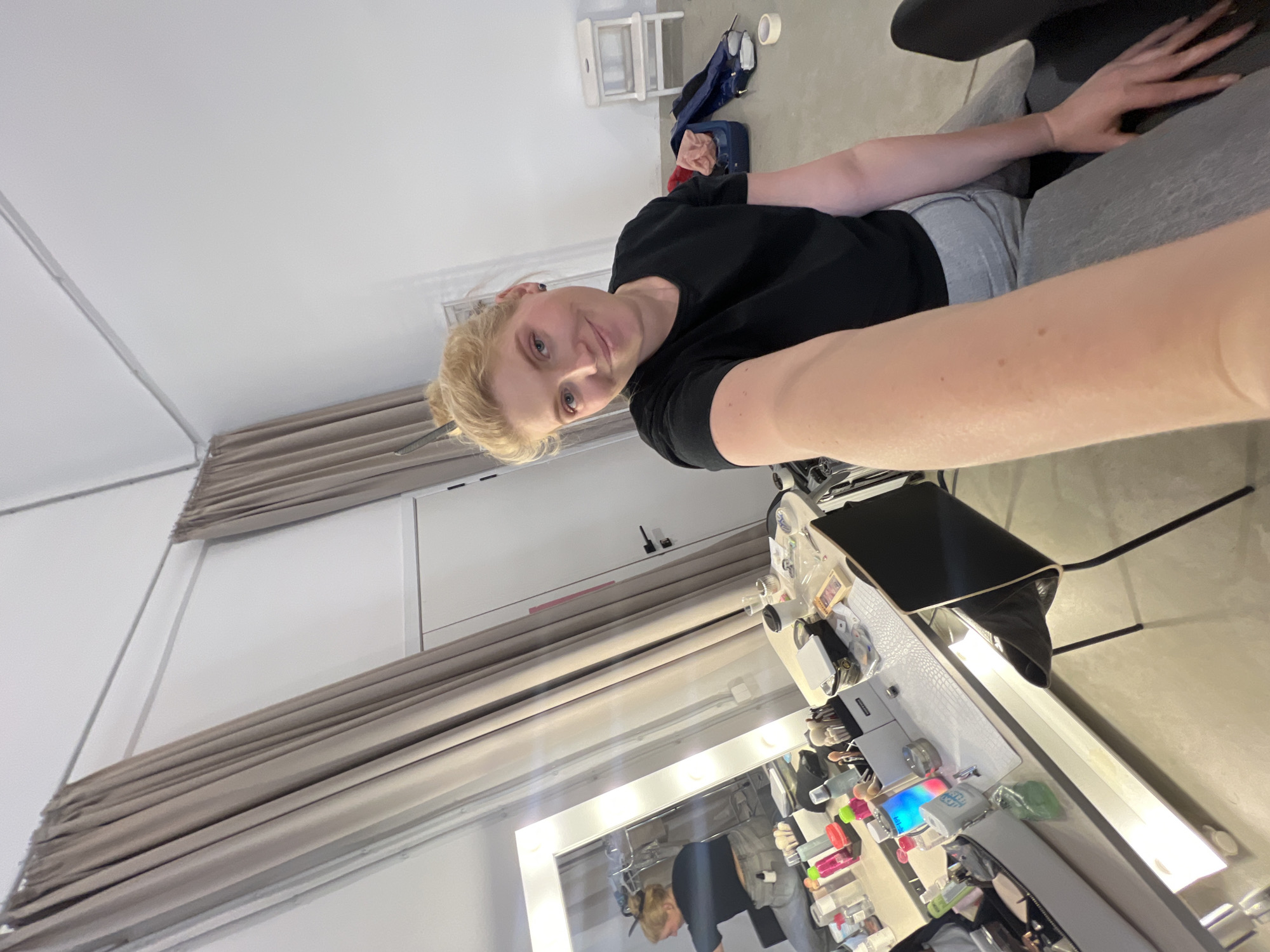 An indoor, medium-shot selfie captures a blonde woman with fair skin, seated and smiling gently towards the camera. She has her hair pulled back and is wearing a black t-shirt and grey bottoms. Her right arm is extended, holding the camera, with her left hand resting on her lap.
She is positioned on a grey upholstered chair or sofa, situated in a brightly lit room. To her right, a large white vanity table with an illuminated mirror dominates the foreground. The table is covered with numerous personal care items, including various bottles, jars, brushes, and cosmetic products, suggesting a makeup or grooming area. A small device emitting colorful light and a black laptop or tablet are also visible on the vanity. A black office chair is partially seen behind the vanity.
The room features white walls and a light-colored floor, possibly concrete or linoleum. Neutral-toned, pleated curtains are drawn across a section of the wall to the left. In the background, behind the woman, a white stool, a blue bag, and a roll of white tape are scattered on the floor, implying a casual or functional setting. The overall atmosphere is relaxed, with the woman appearing content in what seems to be a personal dressing room or a dedicated vanity space, possibly in Khimki, Russia. No visible text is clearly legible in the image.